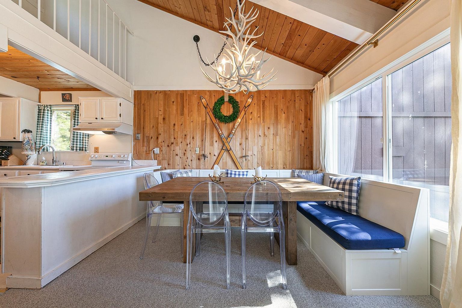This dining area features a rustic wooden table paired with modern, transparent chairs and a built-in bench seating arrangement against a wood-paneled wall. A distinctive antler chandelier hangs above the table, while a decorative wreath and vintage skis add a cozy, mountain-lodge aesthetic. Large windows to the right provide ample natural light, creating an inviting space that flows directly into the adjacent kitchen.