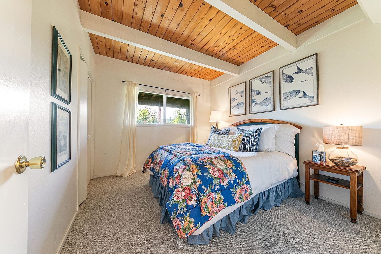 This cozy bedroom features a vaulted wood-paneled ceiling with exposed beams, creating a warm and rustic atmosphere. The room is furnished with a bed dressed in a vibrant floral quilt and a matching blue bed skirt, complemented by a wooden nightstand with a lamp. Three framed fish-themed artworks hang above the headboard, while a window with light curtains allows natural light to brighten the space.
