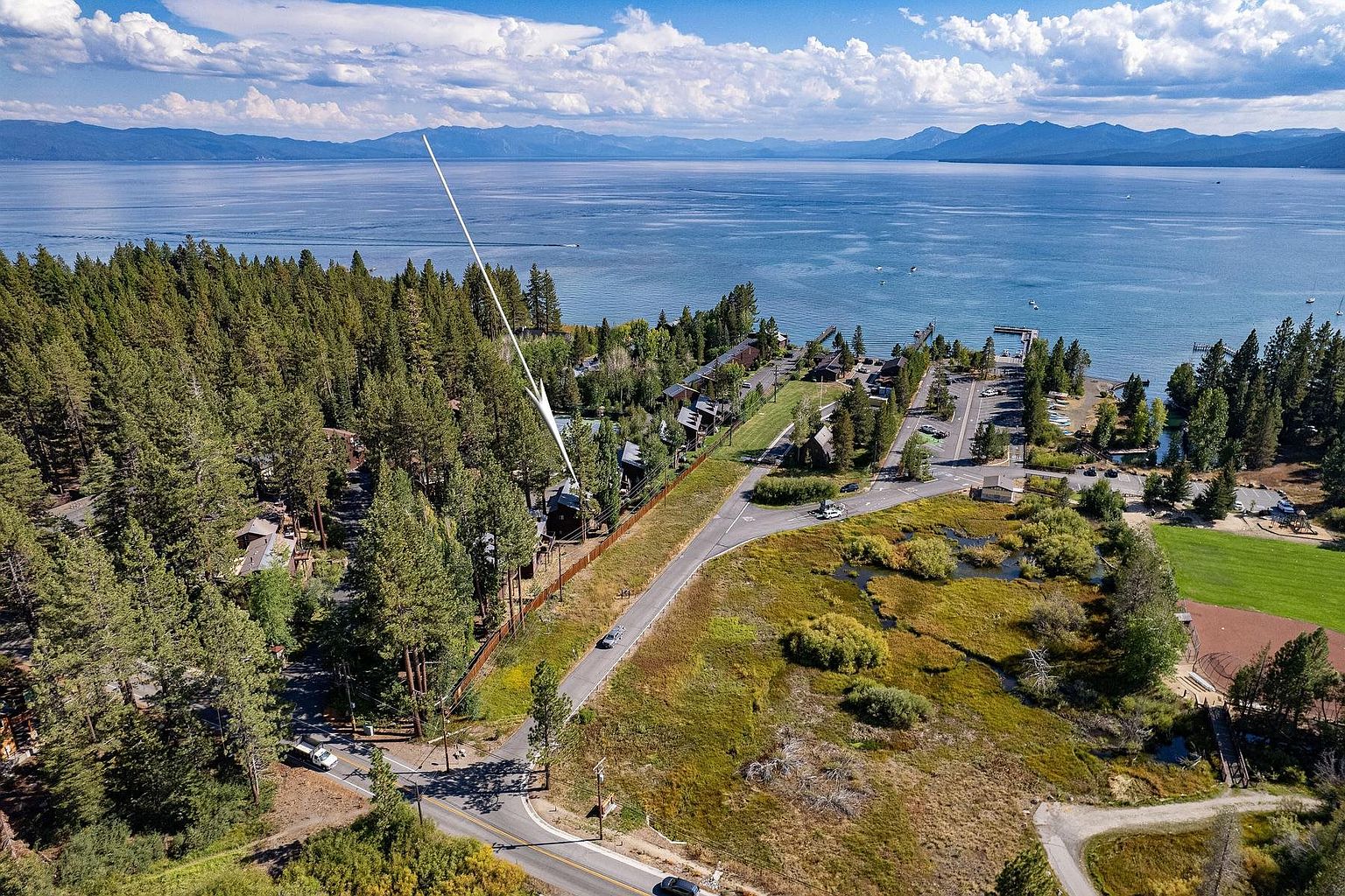 This high-angle aerial view captures a scenic lakeside property nestled within a dense forest, with a white arrow pointing toward a specific cabin. The landscape features a winding road, a grassy marsh area, and a clear view of a large, expansive lake framed by distant mountains under a bright, cloudy sky. The perspective emphasizes the property's proximity to both the natural woodland environment and the waterfront.