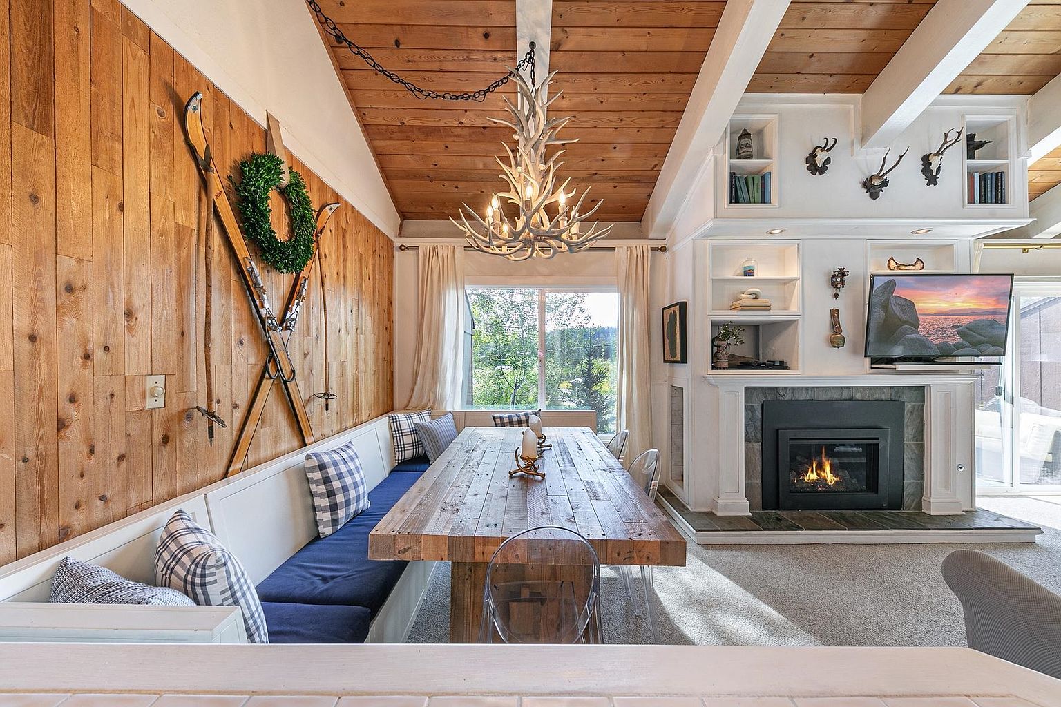 This cozy dining area features a rustic wooden table paired with a built-in bench seating arrangement adorned with blue and white plaid cushions. The space is accented by a prominent antler chandelier, a wood-paneled feature wall decorated with vintage skis, and a fireplace with a stone surround and a mounted television. Large windows allow natural light to fill the room, creating a warm and inviting atmosphere with a distinct mountain-lodge aesthetic.