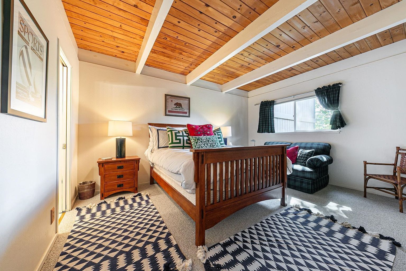 This bedroom features a warm, rustic aesthetic highlighted by a wood-planked ceiling with exposed beams. The room is furnished with a wooden bed frame, a matching nightstand with a lamp, and a plaid-upholstered loveseat positioned near a window. A bold, geometric-patterned rug anchors the space, creating a cozy and inviting atmosphere.