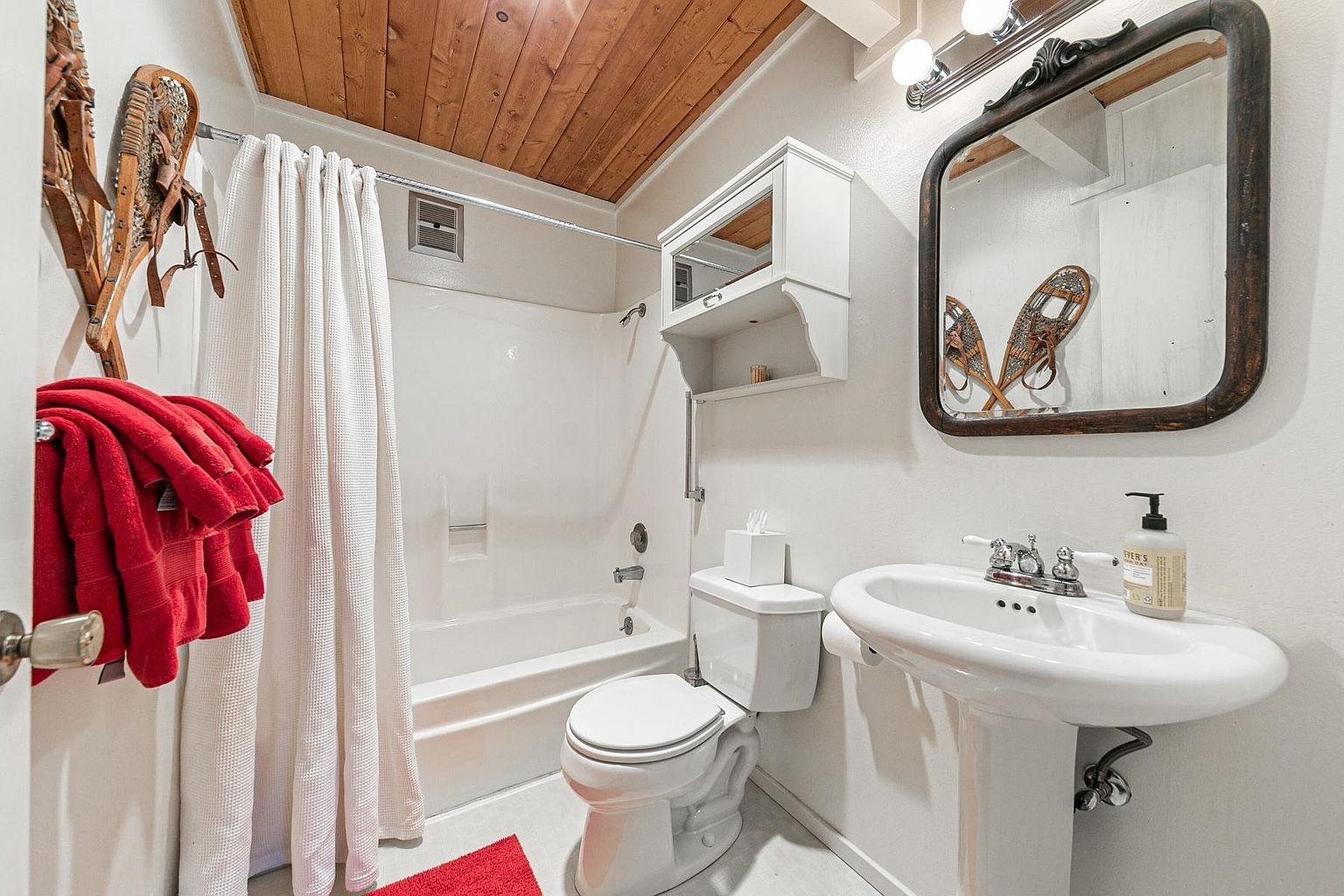 This compact bathroom features a white pedestal sink, a standard toilet, and a shower-tub combination with a white curtain. The space is accented by a rustic wood-paneled ceiling and decorative snowshoes mounted on the wall, creating a cozy, cabin-like aesthetic. A wall-mounted medicine cabinet and a dark-framed mirror complete the functional layout.