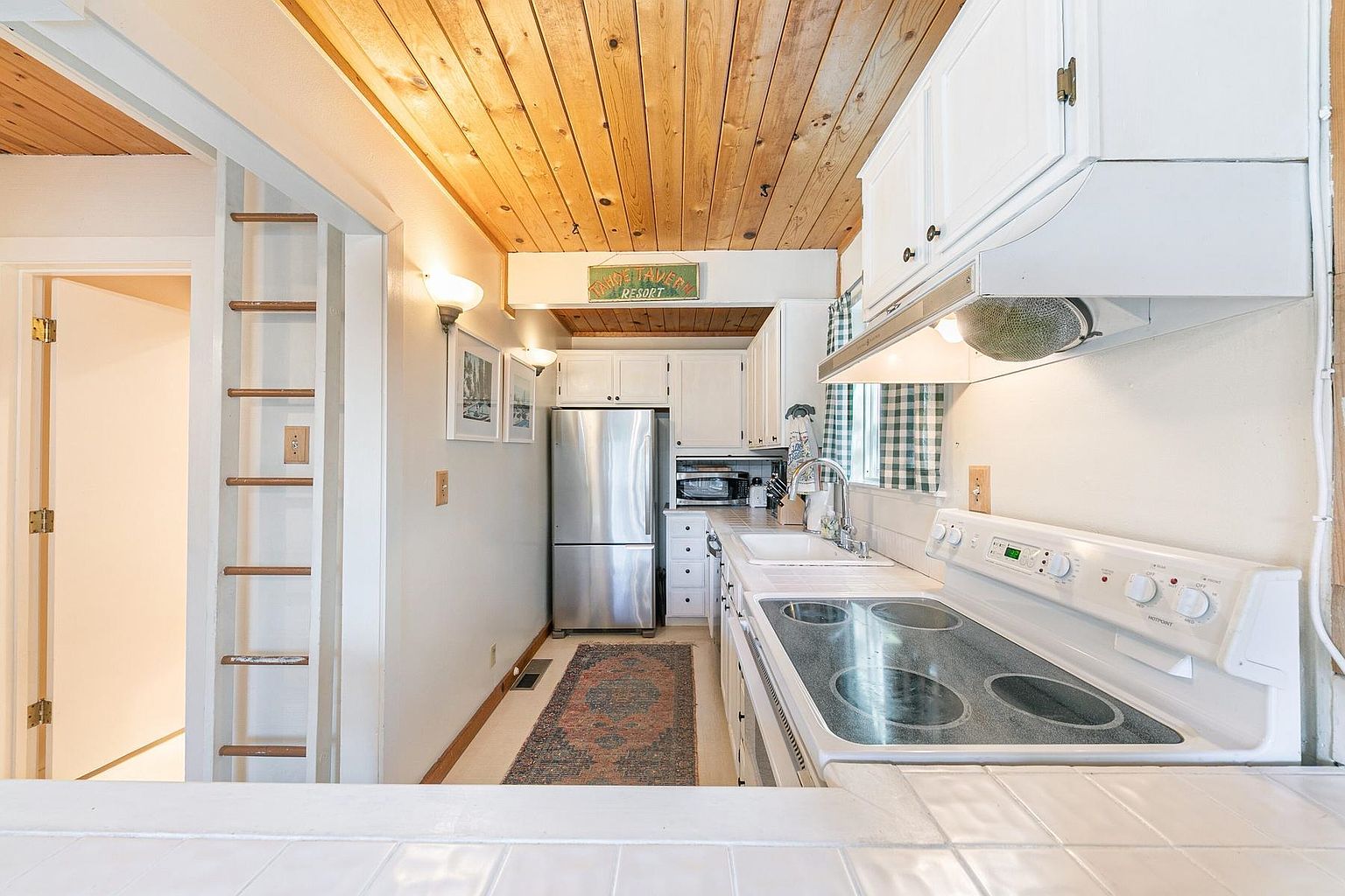 This galley-style kitchen features white cabinetry, a stainless steel refrigerator, and a white electric range. The space is characterized by a warm wood-plank ceiling and a patterned runner rug that adds texture to the floor. A decorative sign above the refrigerator adds a rustic touch, while the perspective from the counter provides a clear view of the functional layout.