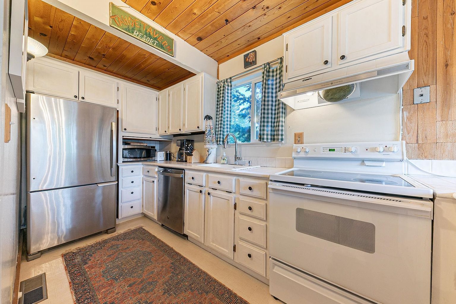This cozy, rustic-style kitchen features white cabinetry paired with a warm wood-paneled ceiling and wood-paneled walls. The space is equipped with a stainless steel refrigerator, a white electric range, and a dishwasher, all arranged in a functional galley layout. A patterned area rug adds a touch of color to the floor, while a window above the sink brings in natural light, creating a welcoming and homey atmosphere.