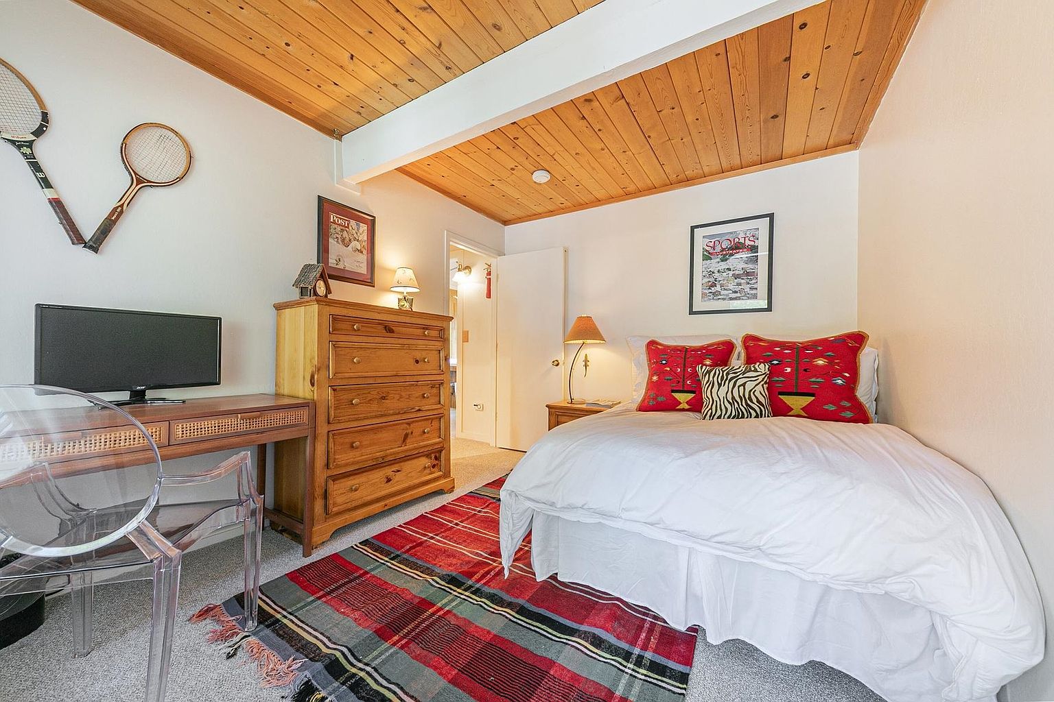 This cozy guest bedroom features a warm, rustic aesthetic with a light wood-paneled ceiling and a plaid area rug. The room is furnished with a bed adorned with vibrant red patterned pillows, a wooden dresser, and a desk with a transparent chair. A framed sports print hangs above the bed, while vintage tennis rackets are mounted on the wall, creating a charming, lodge-inspired atmosphere.