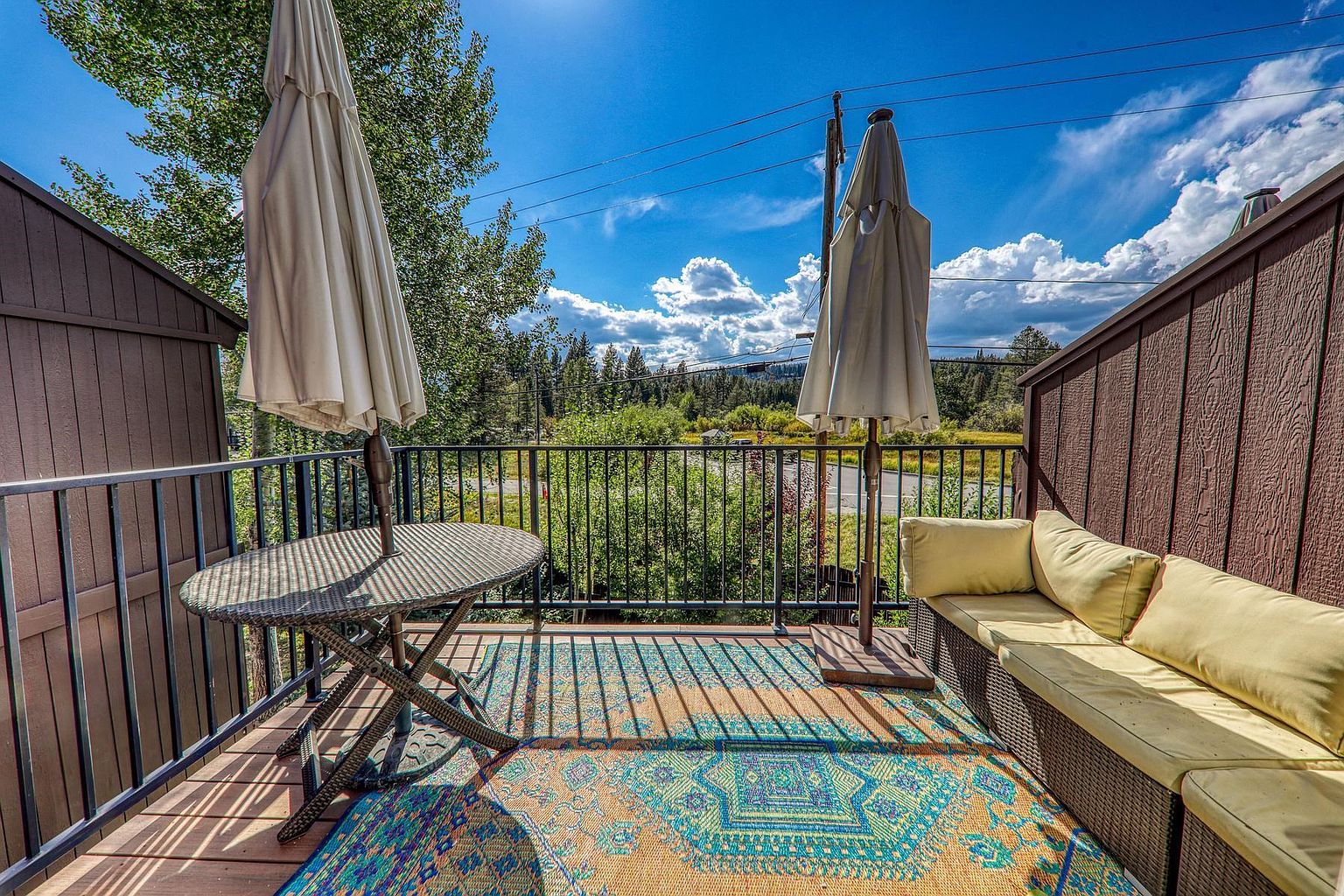 This elevated balcony features a cozy outdoor seating area with a wicker sofa and a matching round table, all set upon a vibrant, patterned area rug. The space is framed by a black metal railing and wooden privacy walls, offering a clear view of the surrounding trees and distant mountains under a bright, sunny sky. Two closed patio umbrellas provide a sense of relaxation, making this an inviting spot for outdoor leisure.