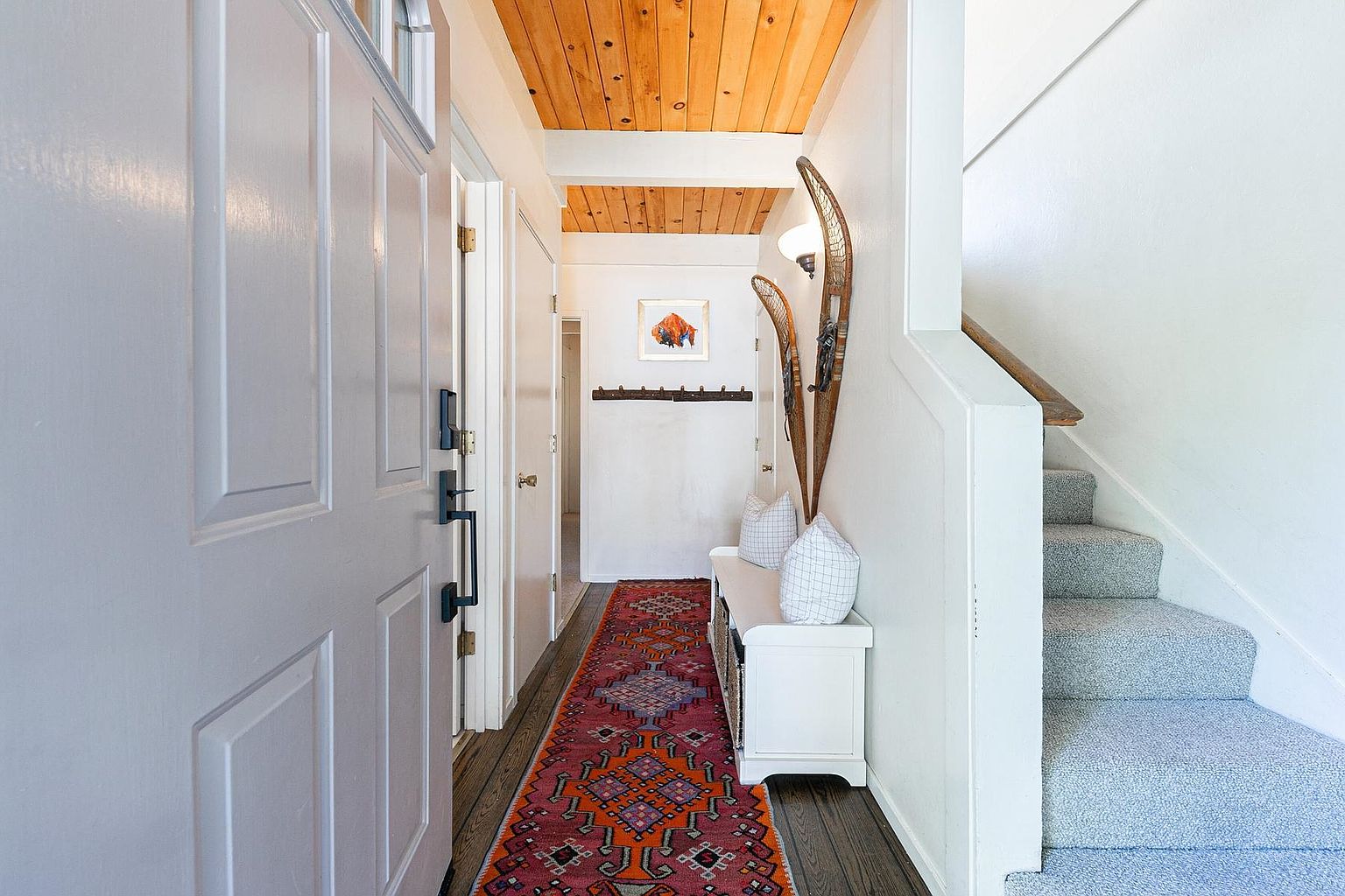 This inviting entryway features a warm, wood-planked ceiling and a vibrant, patterned runner that leads down the hallway. To the right, a white bench with patterned pillows sits beneath decorative snowshoes mounted on the wall, while a carpeted staircase ascends to the upper level. The perspective is from the front door looking inward, showcasing a bright, cozy transition space with a clean, rustic-inspired aesthetic.