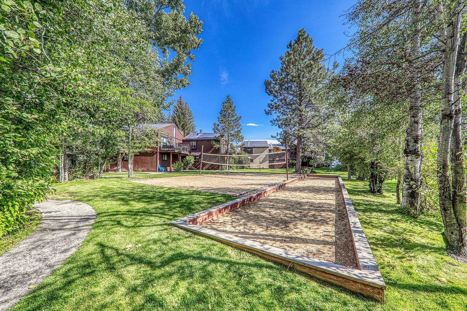 This outdoor community space features a sand volleyball court and a separate bocce ball court framed by wooden borders. The area is surrounded by lush green grass and mature trees, with multi-story brown wood-sided residential buildings visible in the background. The perspective is a wide-angle ground-level shot that captures the recreational amenities set within a peaceful, wooded residential environment.