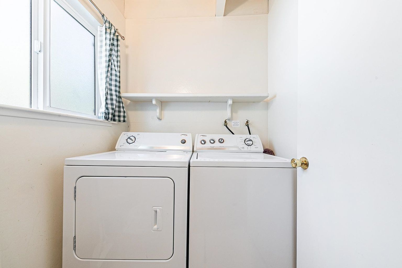 This functional laundry room features a side-by-side washer and dryer set positioned beneath a simple white wall-mounted shelf. A window with a checkered curtain provides natural light to the space, which is finished with neutral-toned walls. The room is compact and utilitarian, offering a straightforward layout for household chores.