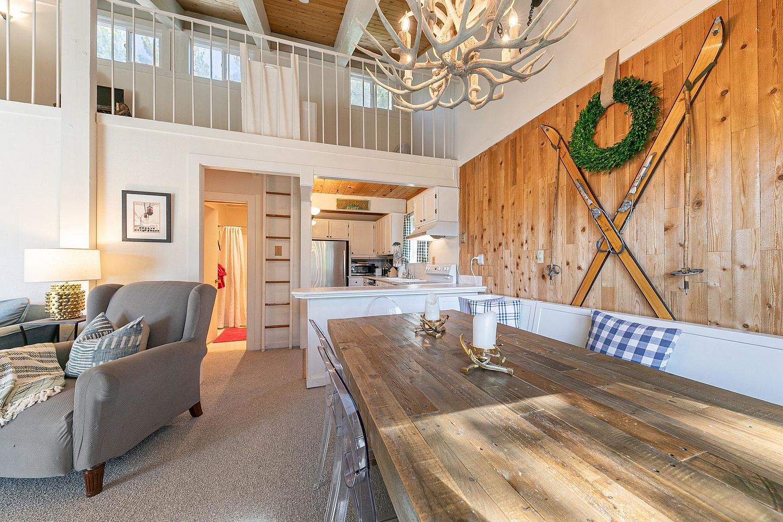 This cozy, rustic-inspired dining area features a large, reclaimed wood table paired with transparent chairs, set against a warm wood-paneled wall decorated with vintage skis and a wreath. The space is illuminated by a striking antler chandelier and flows into an open-concept kitchen with white cabinetry and stainless steel appliances. A lofted area with a white railing overlooks the room, adding architectural interest and a sense of vertical openness to the cabin-style interior.