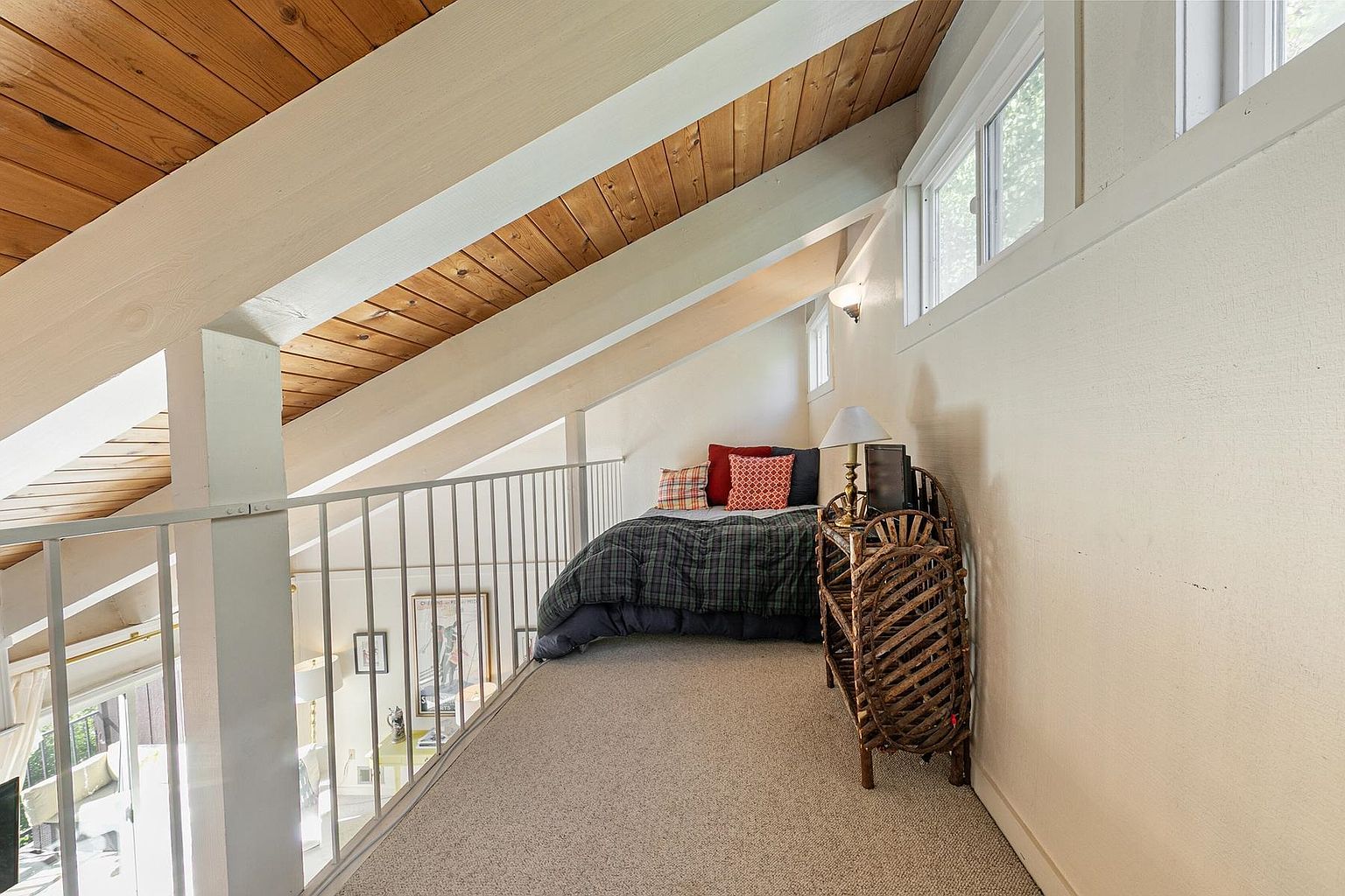 This cozy loft space features a sloped wood-paneled ceiling and a built-in bed tucked into the eaves. A white metal railing overlooks the lower level, while a unique, curved wicker shelving unit stands beside the bed. The room is carpeted and illuminated by natural light from a window, creating a warm and inviting nook.