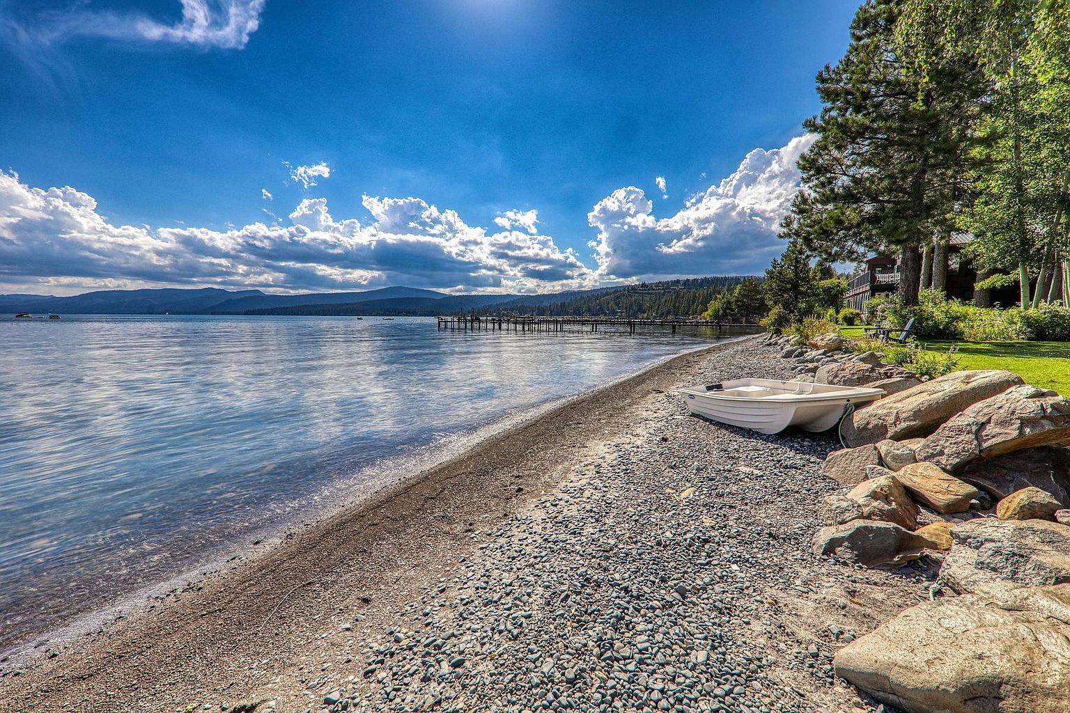 This scenic waterfront view captures a tranquil pebble beach along a large lake, framed by a lush green lawn and mature pine trees. A small white rowboat rests on the shore near a cluster of large boulders, while a long wooden pier extends into the calm water toward distant mountains. The bright blue sky with scattered clouds creates a peaceful and inviting atmosphere, highlighting the property's direct access to the water.