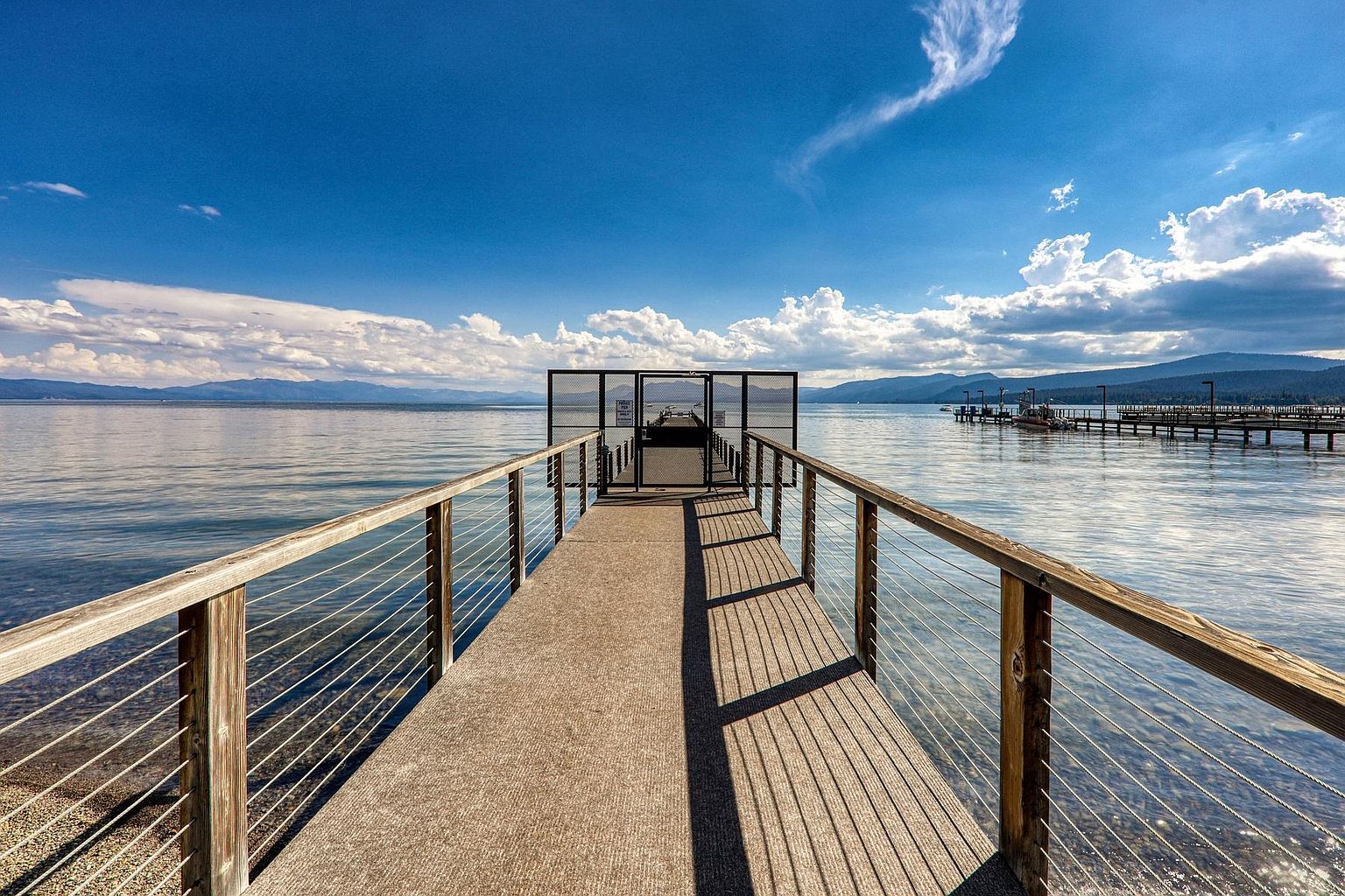 This perspective captures a long, wooden pier extending out over a calm, expansive lake under a bright blue sky with scattered clouds. The walkway features a sturdy railing with tensioned cable infill and leads to a gated entrance, beyond which another pier is visible in the distance. The scene conveys a serene, recreational atmosphere, highlighting the property's direct access to waterfront amenities.