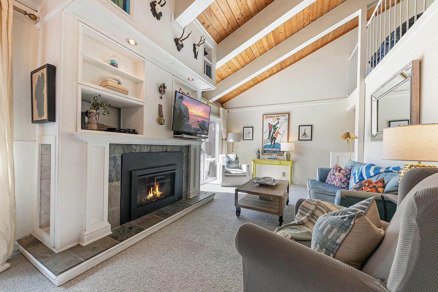 This cozy living room features a vaulted wood-paneled ceiling and a prominent stone-clad fireplace with a mounted television above the mantel. The space is furnished with comfortable seating, including a grey armchair and a blue sofa, arranged around a wooden coffee table. A bright yellow console table sits against the back wall, accented by a vintage-style travel poster, creating a warm and inviting atmosphere.