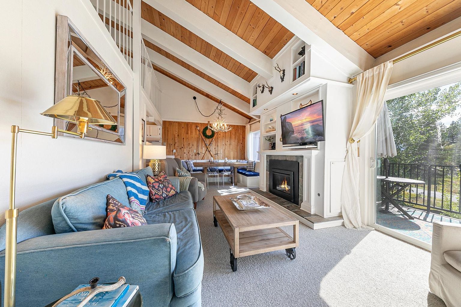This inviting living room features a vaulted ceiling with exposed wood beams and a cozy, cabin-inspired aesthetic. A blue sofa sits opposite a fireplace with a stone-clad chimney breast and a mounted television, while a sliding glass door provides access to an outdoor balcony. The space is accented by rustic decor, including antler motifs and a light wood coffee table, creating a warm and comfortable atmosphere.