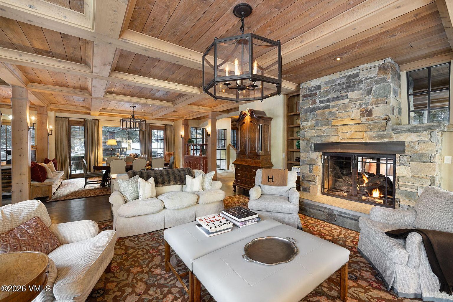 This is a warm and inviting living room featuring a stone fireplace, wood-paneled ceiling, and comfortable seating. The room is decorated in neutral tones with pops of color in the rug and accessories. The overall impression is one of rustic elegance and comfort, perfect for relaxing and entertaining.