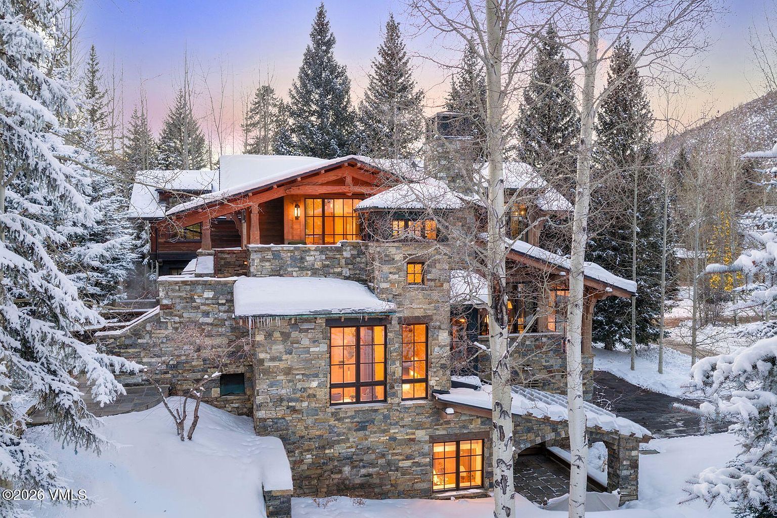 This is a front exterior view of a luxurious multi-story stone house in a snowy, wooded setting. The house features a combination of stone and wood construction, with large windows that glow warmly, suggesting a cozy interior. The surrounding landscape is covered in snow, with evergreen trees and bare birch trees adding to the wintery ambiance.