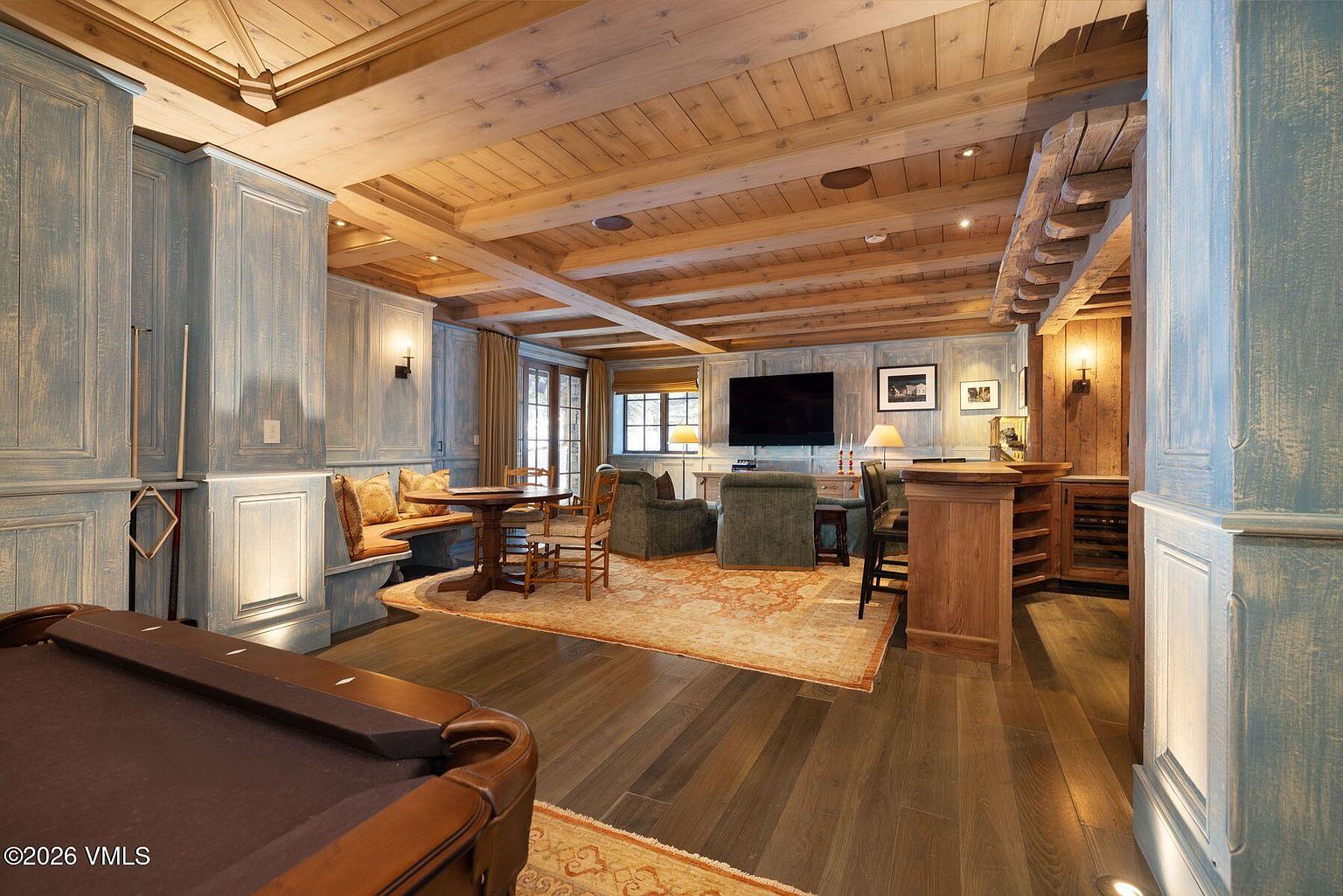 This inviting living room features a blend of rustic and refined elements. The room is anchored by a pool table and includes a seating area with comfortable armchairs, a built-in bench, and a small dining table. The wooden ceiling beams and paneling add warmth, while the area rug ties the space together, creating a cozy and sophisticated atmosphere.