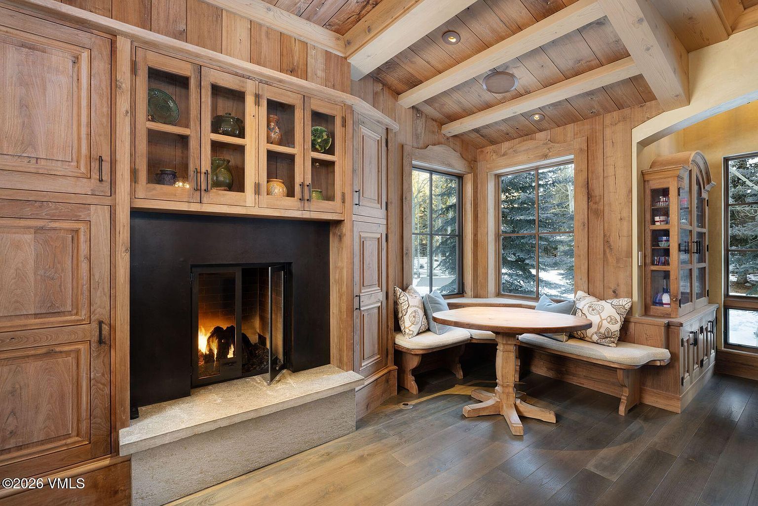 This inviting dining area features rustic wood paneling on the walls and ceiling, creating a warm and cozy atmosphere. A round wooden table is surrounded by built-in bench seating with patterned pillows, nestled between two windows that offer natural light and views of the outdoors. A fireplace with glass-front doors and built-in cabinetry adds to the room's charm and functionality.