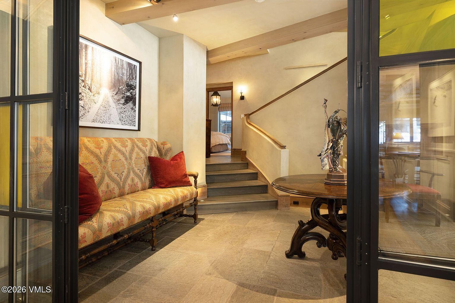 This interior shot showcases a hallway with stone flooring, a staircase leading to an upper level, and a decorative sofa with red pillows. A round wooden table with a statue sits near a window, adding a touch of elegance. The overall style is warm and inviting, with a blend of rustic and sophisticated elements.
