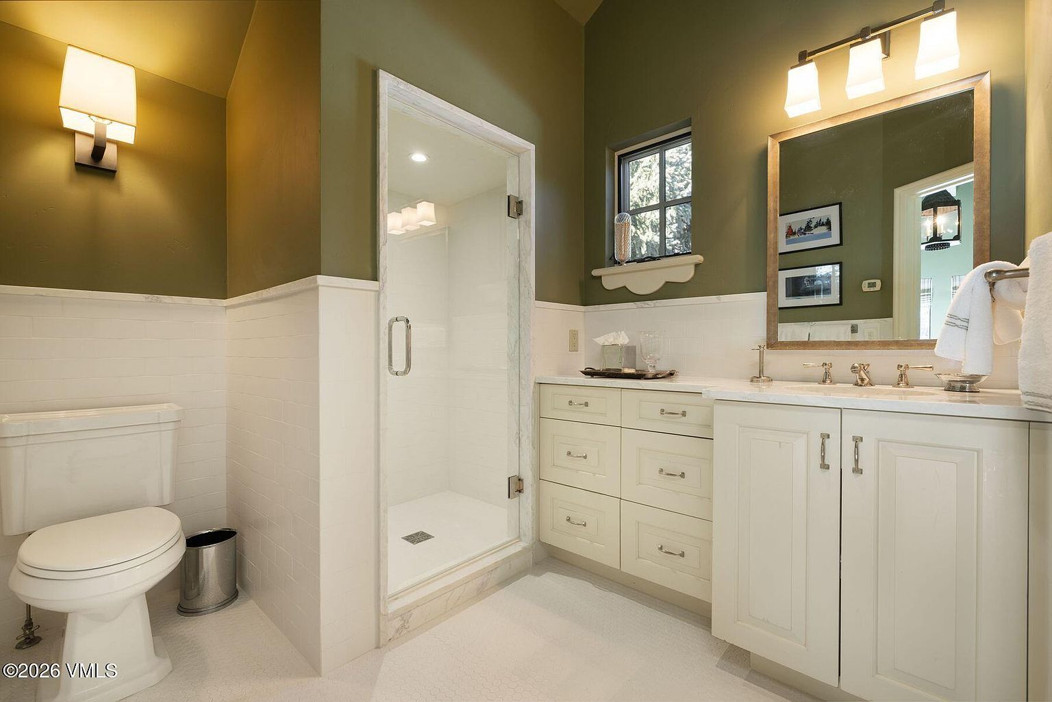 This is a well-lit bathroom featuring a toilet, a glass-enclosed shower, and a vanity with drawers and cabinets. The walls are painted in a two-tone style, with a lower section of white subway tiles and an upper section of olive green paint. The overall impression is clean and functional, with a touch of traditional style.