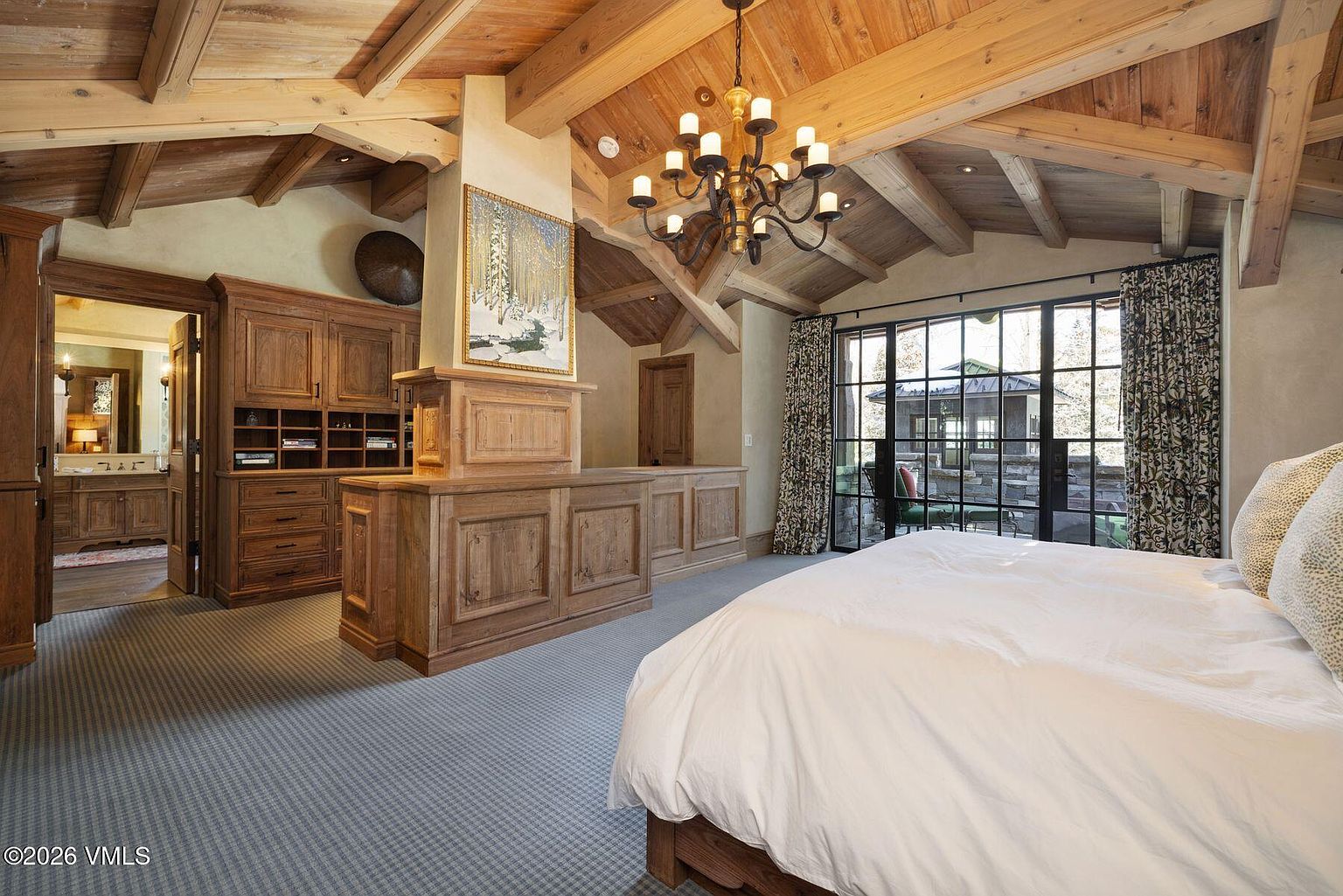 This is a primary bedroom featuring a wooden beamed ceiling, a large bed with white linens, and a built-in wooden dresser with a painting above it. The room has a rustic yet luxurious feel, with a large window offering natural light and a view to the outside. A chandelier hangs from the ceiling, adding to the room's elegance.