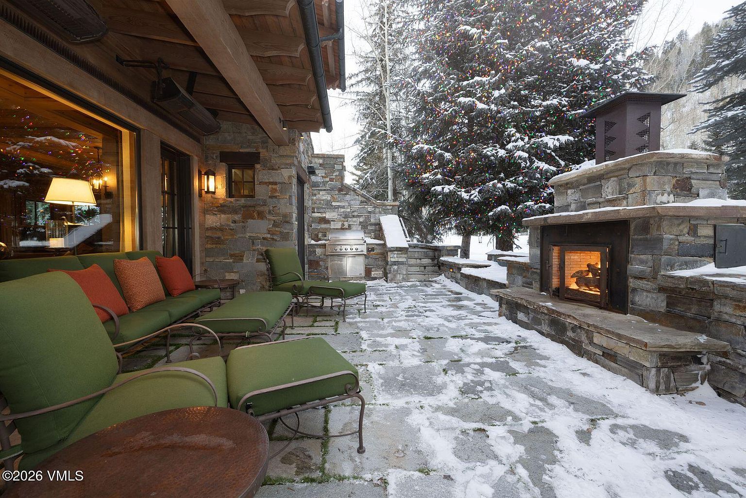 This image showcases a luxurious outdoor patio area, complete with comfortable green cushioned seating, an outdoor fireplace constructed of stone, and a built-in grill. The patio is partially covered in snow, suggesting a winter setting, and a decorated evergreen tree adds a festive touch. The stone construction and warm lighting create an inviting and upscale atmosphere.