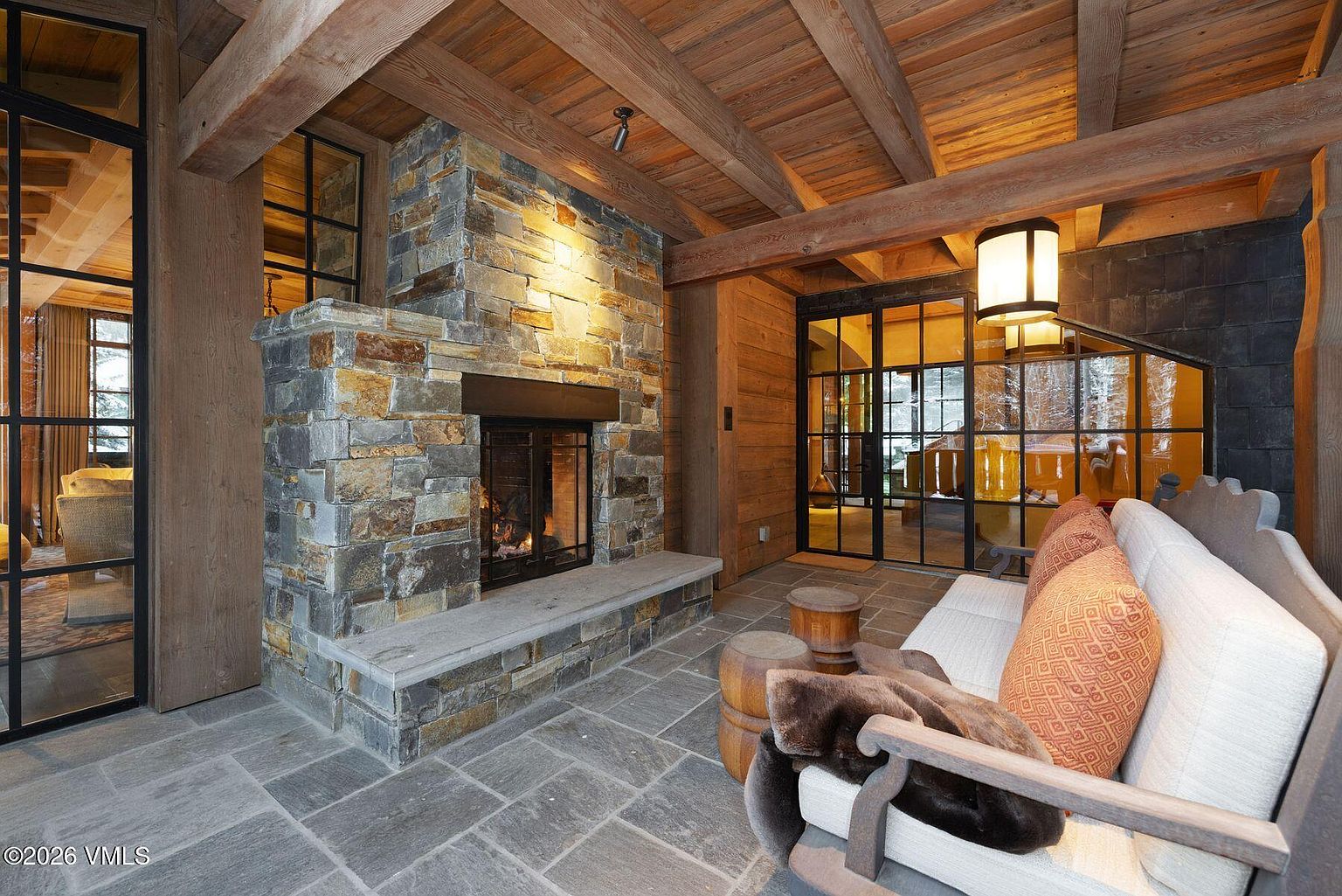 This inviting living room features a stone fireplace as its focal point, complemented by exposed wooden beams on the ceiling. Large windows with black frames offer views to the outside, enhancing the room's natural light. A comfortable seating area with plush cushions and a fur throw creates a cozy and luxurious atmosphere.