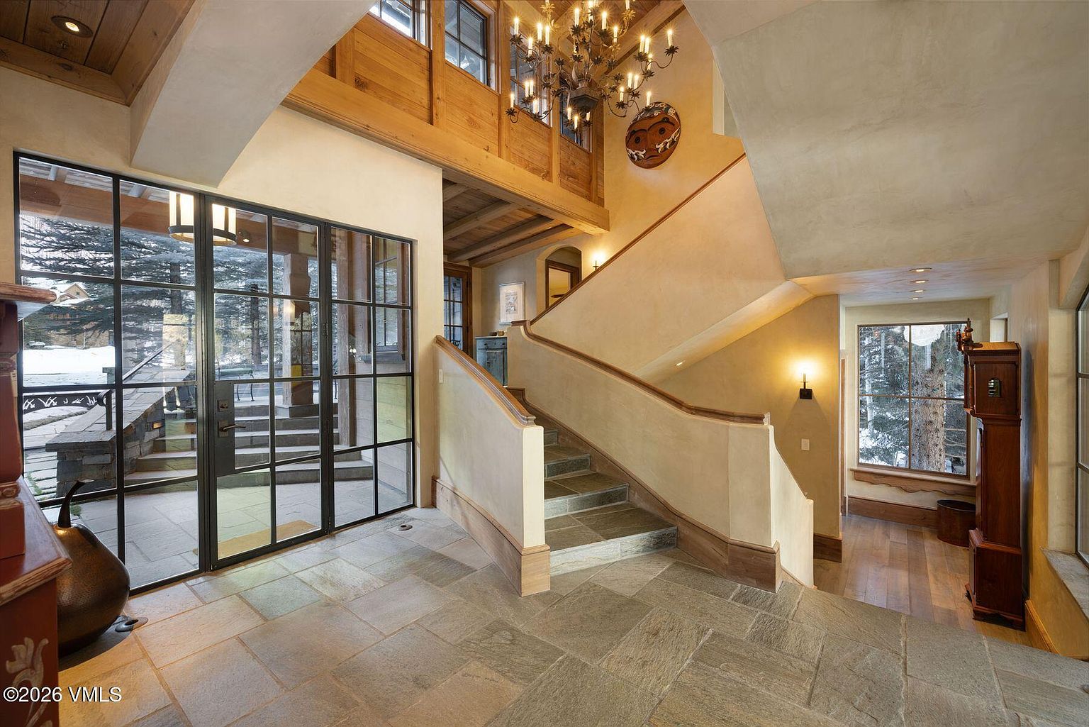 This interior shot showcases a grand entryway featuring stone flooring and a staircase with a light-colored banister. A large window with black frames offers a view of the snowy outdoors, while an ornate chandelier hangs above. The space exudes a luxurious and inviting atmosphere, blending rustic charm with modern design elements.