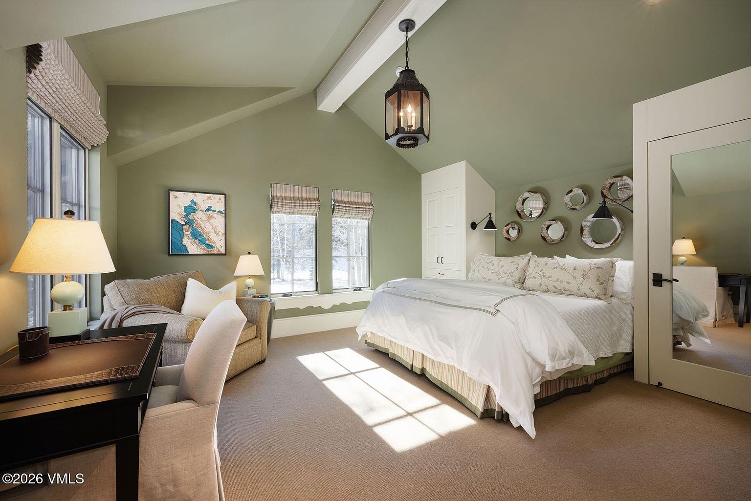 This primary bedroom features a calming green color scheme, complemented by natural light streaming through the windows. The room includes a king-size bed with white linens, a seating area with a sofa, and a built-in wardrobe. Decorative mirrors adorn the wall above the bed, adding a touch of elegance.