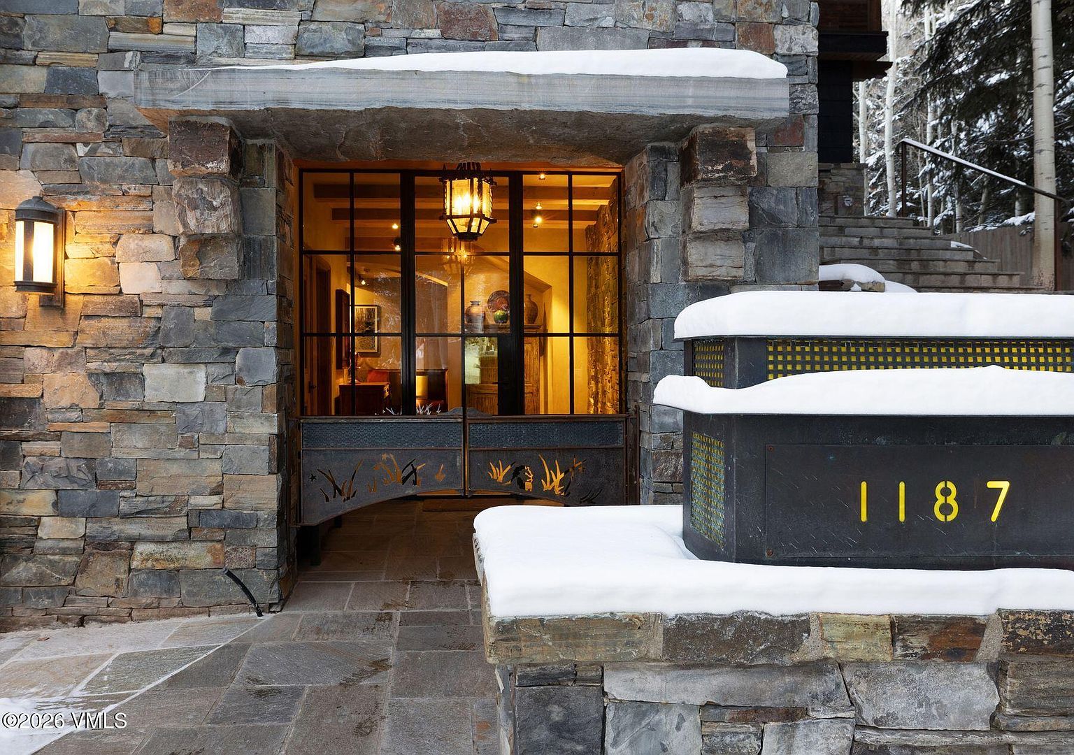 This image showcases the grand entryway of a stone-clad residence, featuring a custom metal gate and a prominent address marker illuminated with yellow numbers. The entrance is framed by substantial stone pillars and a lintel, with snow accents adding a touch of winter charm. The overall impression is one of rustic elegance and solid construction.