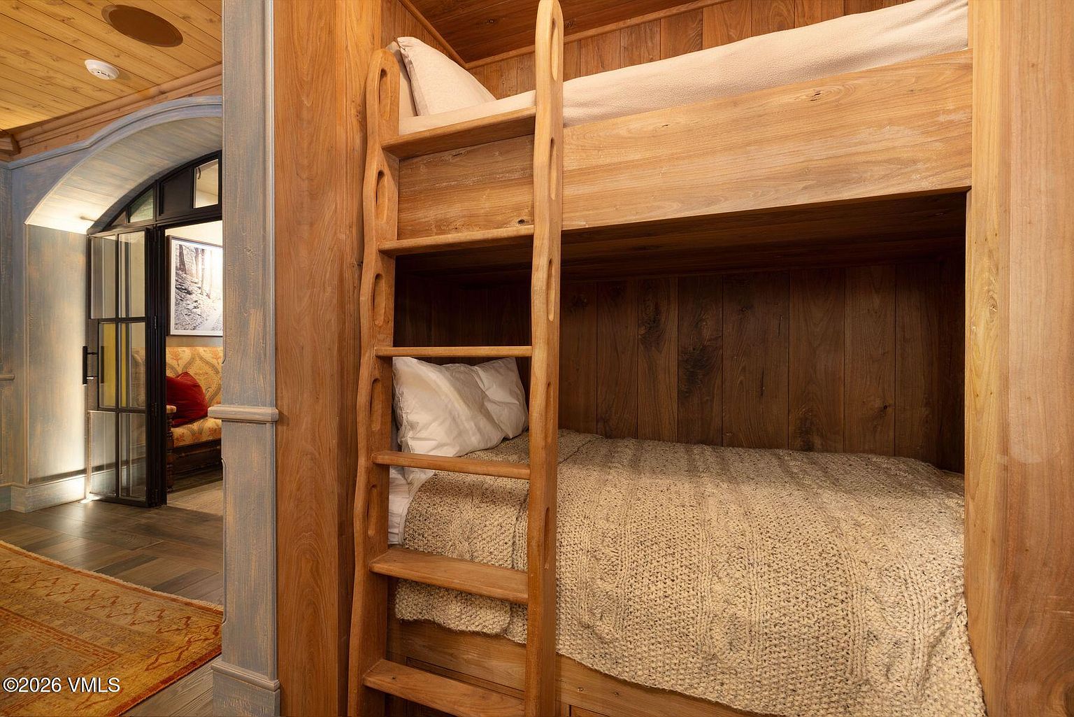 This image showcases a cozy bedroom featuring custom-built wooden bunk beds. The room exudes a rustic charm with its wood paneling and warm lighting. A glimpse into an adjacent room reveals a comfortable seating area, enhancing the inviting atmosphere of the space.