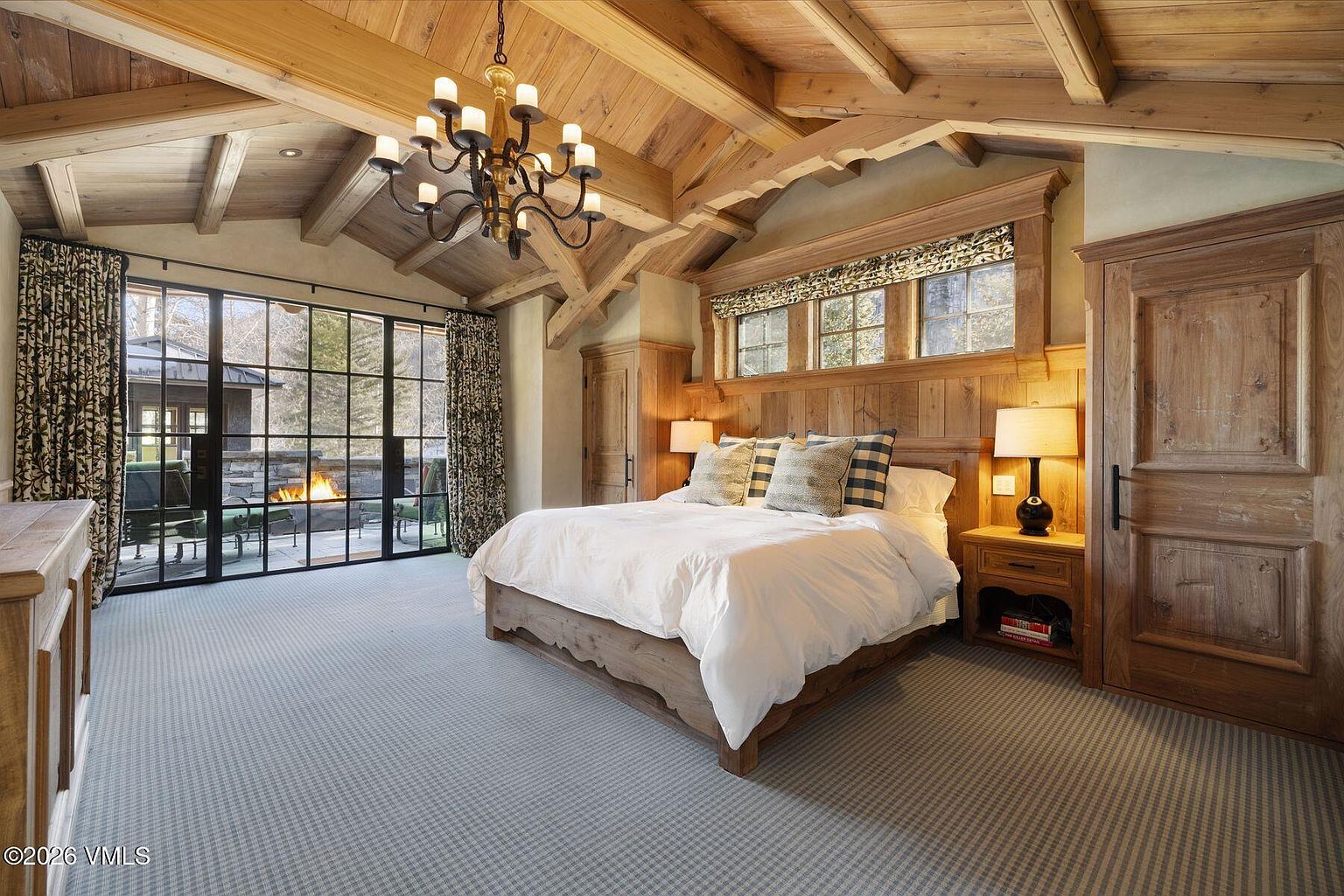 This is a primary bedroom featuring a wooden vaulted ceiling with exposed beams and a large window wall with black frames that leads to an outdoor patio with a fireplace. The room is furnished with a wooden bed frame, white linens, and a wooden nightstand with a lamp. The overall style is rustic and luxurious, creating a warm and inviting atmosphere.
