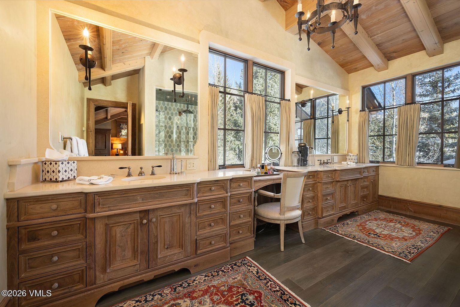 This is a luxurious primary bathroom featuring dual wooden vanities with marble countertops, large mirrors, and natural light streaming through multiple windows. The bathroom has a rustic-chic aesthetic with exposed wooden beams on the ceiling and elegant rugs on the dark wood floor, creating a warm and inviting atmosphere. The perspective is from the doorway, showcasing the spacious layout and high-end finishes.
