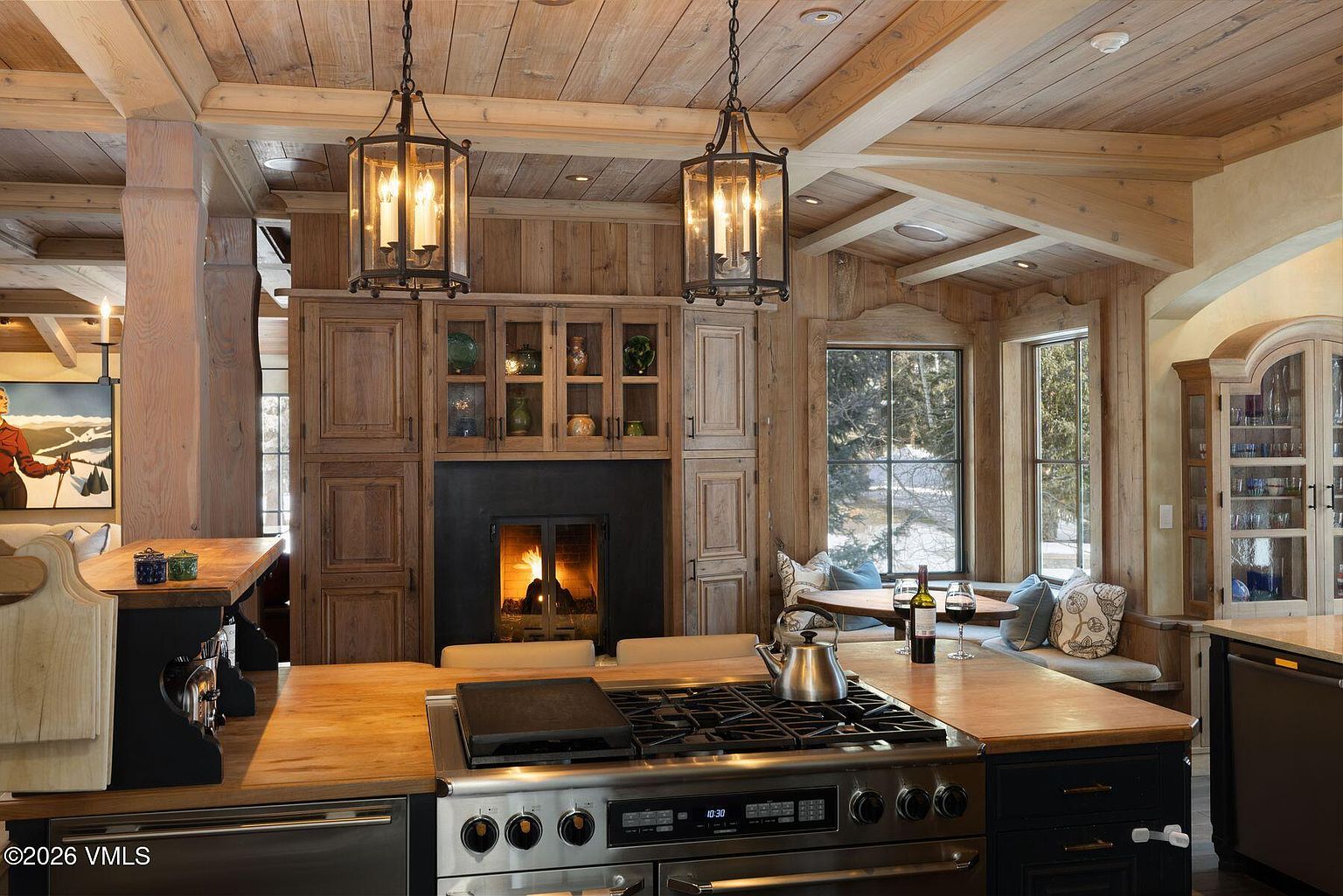 This is a warm and inviting kitchen featuring wood-paneled walls and ceiling, creating a rustic yet elegant atmosphere. The kitchen boasts a large stainless steel range with a wooden countertop, complemented by hanging lantern-style light fixtures. A cozy breakfast nook with windows offers a view of the outdoors, enhancing the kitchen's charm and appeal.