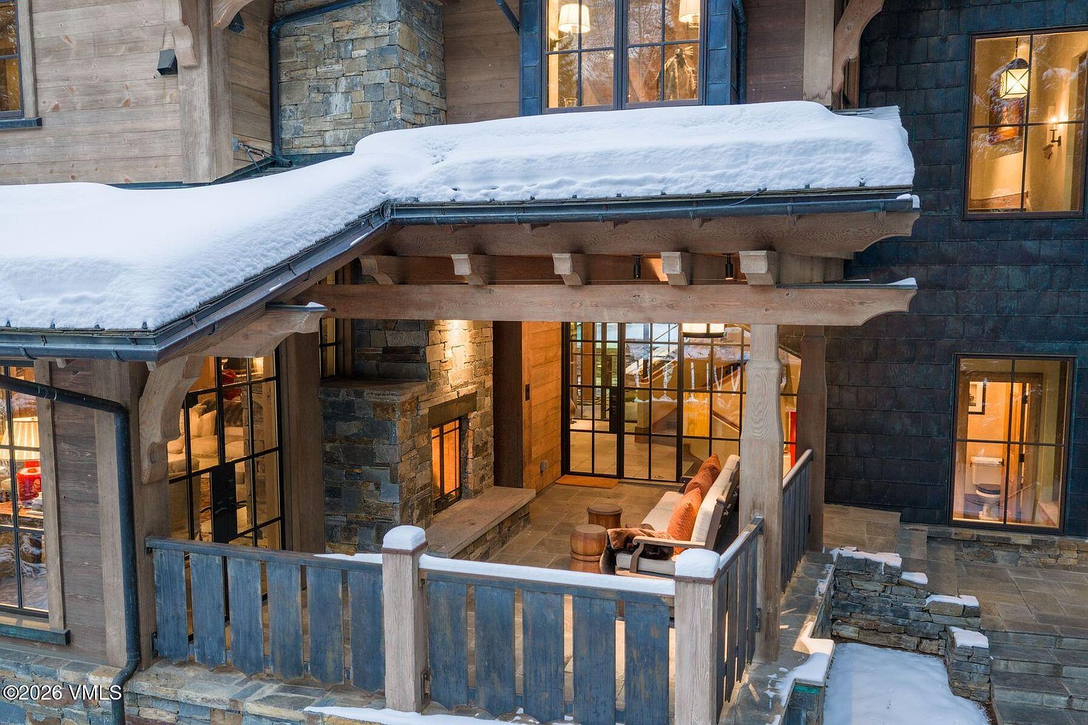 This image showcases a beautifully designed outdoor patio area, featuring a stone fireplace, comfortable seating, and a rustic wooden railing. The patio is partially covered by a snow-laden roof, creating a cozy and inviting atmosphere. The architectural style blends natural materials with modern design elements, making it an ideal space for relaxation and entertainment.