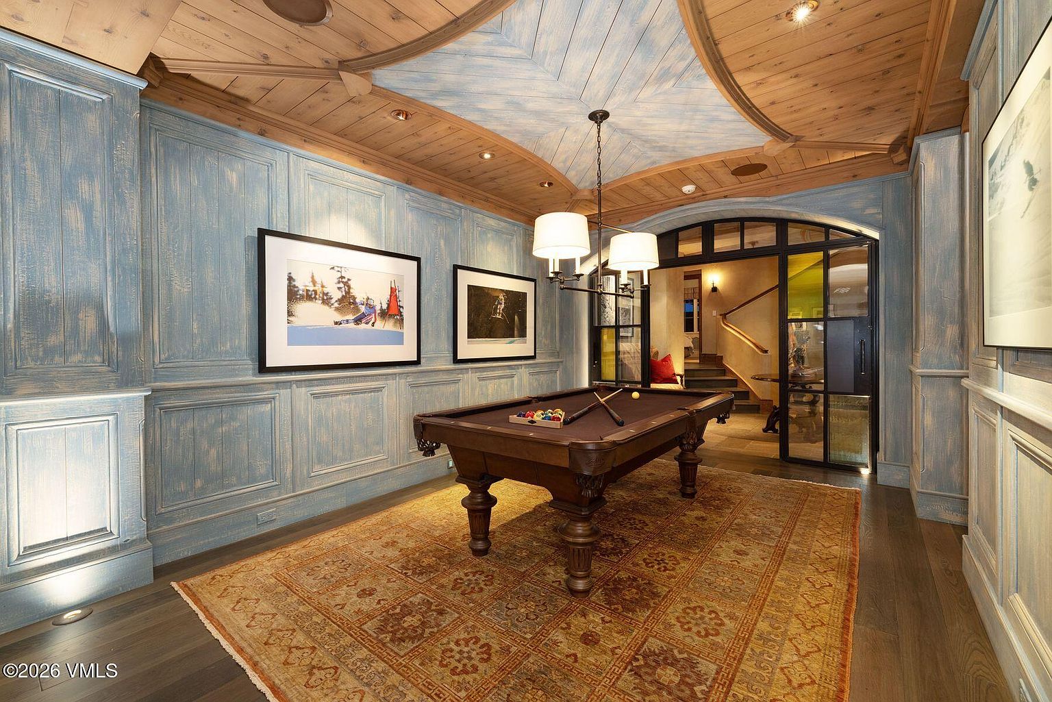 This interior shot showcases a sophisticated living room featuring a pool table as a central element. The room is adorned with elegant wood paneling in a light blue hue, complemented by a patterned rug and framed artwork. A unique wooden ceiling design and a stylish chandelier add to the room's luxurious and inviting atmosphere.