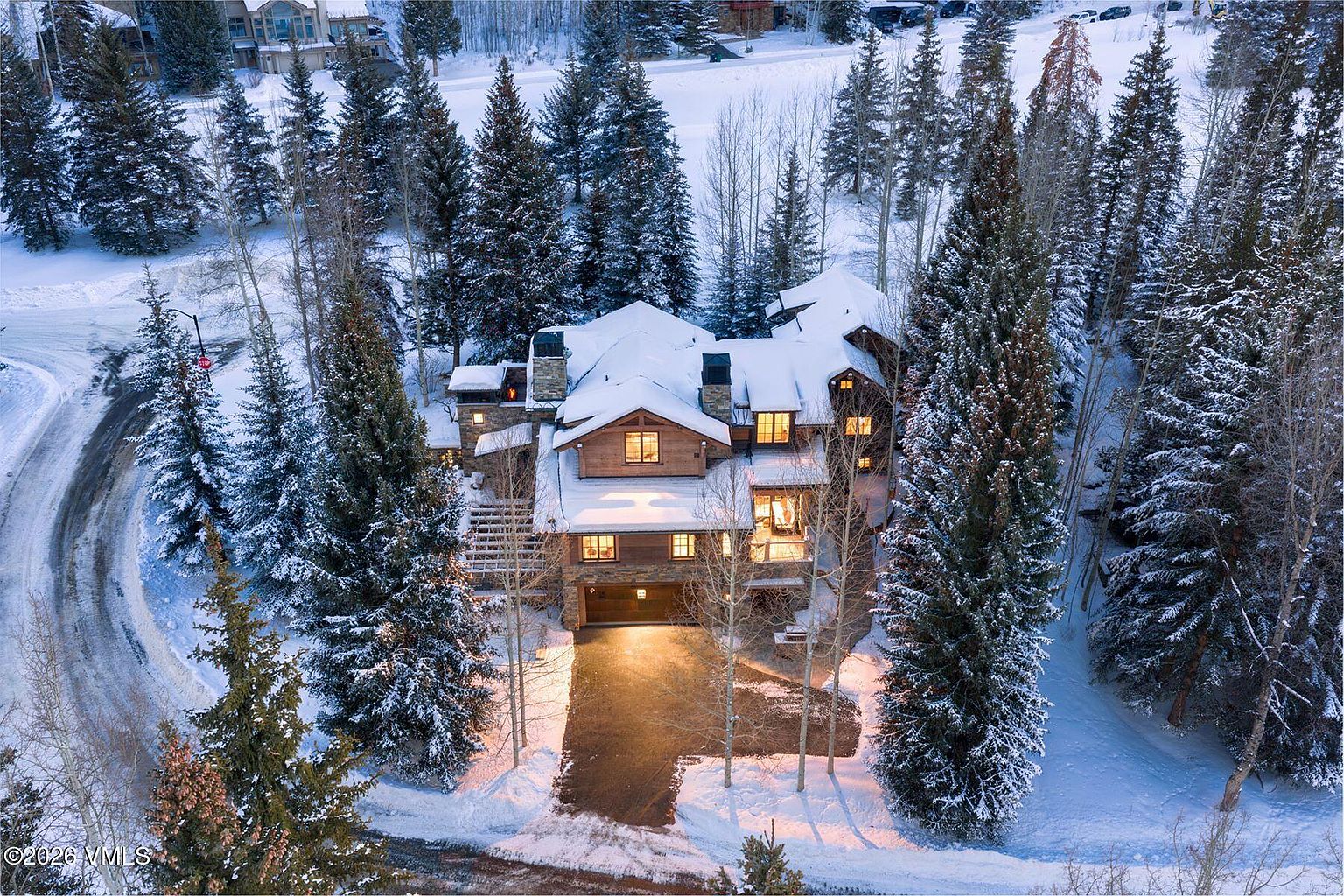 This aerial view showcases a luxurious mountain home nestled among snow-covered evergreen trees. The multi-story residence features a combination of stone and wood exterior, with a snow-covered roof and multiple chimneys. Warm light emanates from the windows, creating an inviting ambiance in the winter landscape.