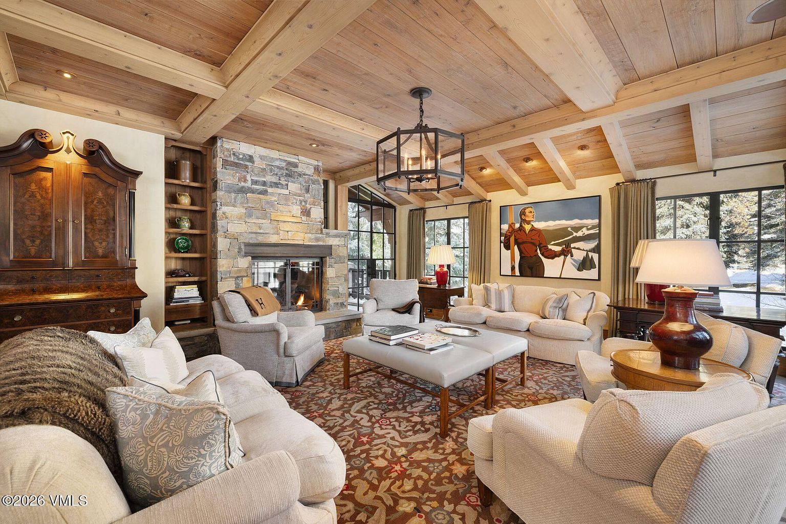 This is a warm and inviting living room featuring a stone fireplace, wood-beamed ceilings, and comfortable seating arrangements. The room is decorated with a mix of traditional and rustic elements, including a large armoire, a patterned rug, and a painting of a skier. Large windows provide natural light and views of the outdoors, creating a cozy and luxurious atmosphere.