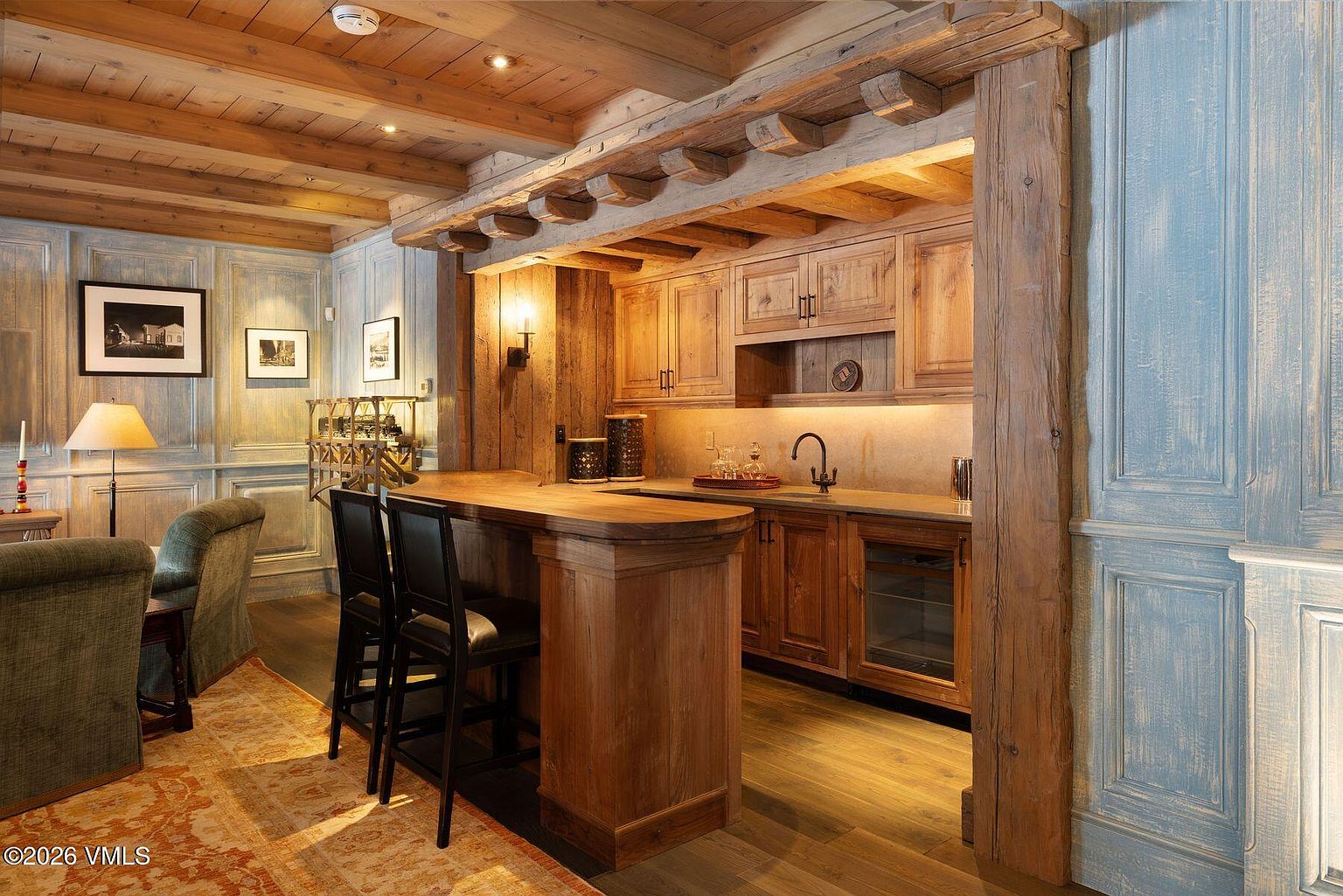 This interior shot showcases a rustic kitchen and bar area with warm wood cabinetry, countertops, and ceiling beams. The space features a bar counter with stools, a sink area, and built-in appliances, all illuminated by warm lighting. The overall impression is cozy and inviting, with a blend of rustic charm and functional design.