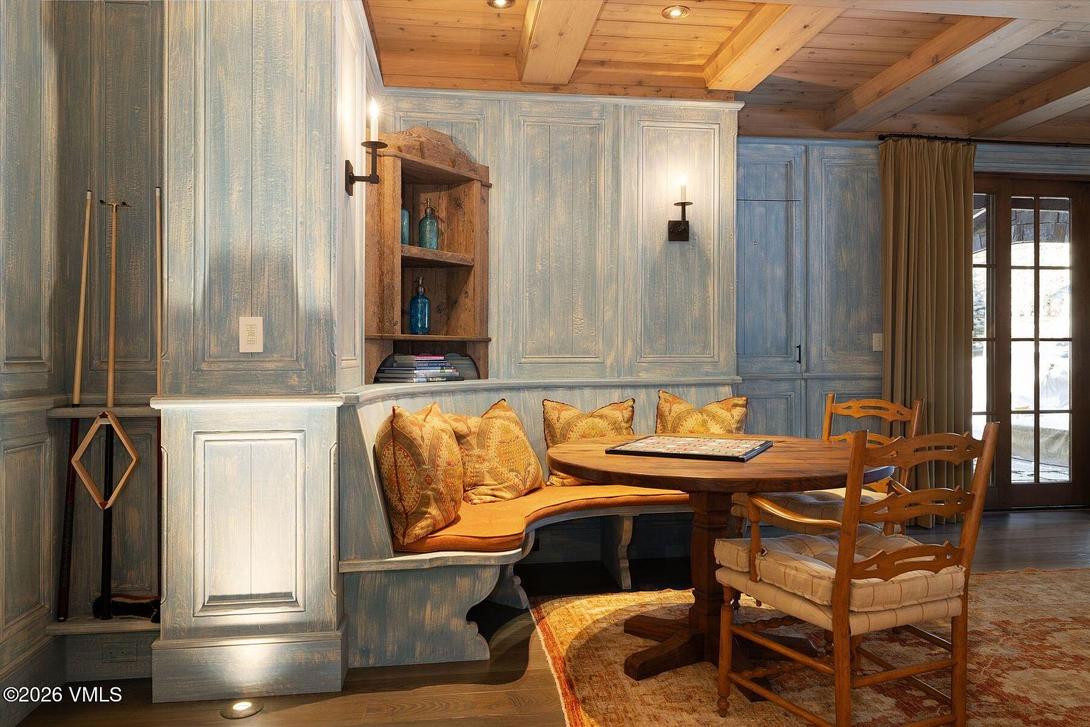 This is a dining area featuring a built-in curved bench with patterned pillows and a round wooden table with chairs. The walls are paneled in a distressed blue finish, and the ceiling has exposed wooden beams. A wooden shelf unit and sconces add to the room's cozy and rustic charm, while a glass door provides natural light.