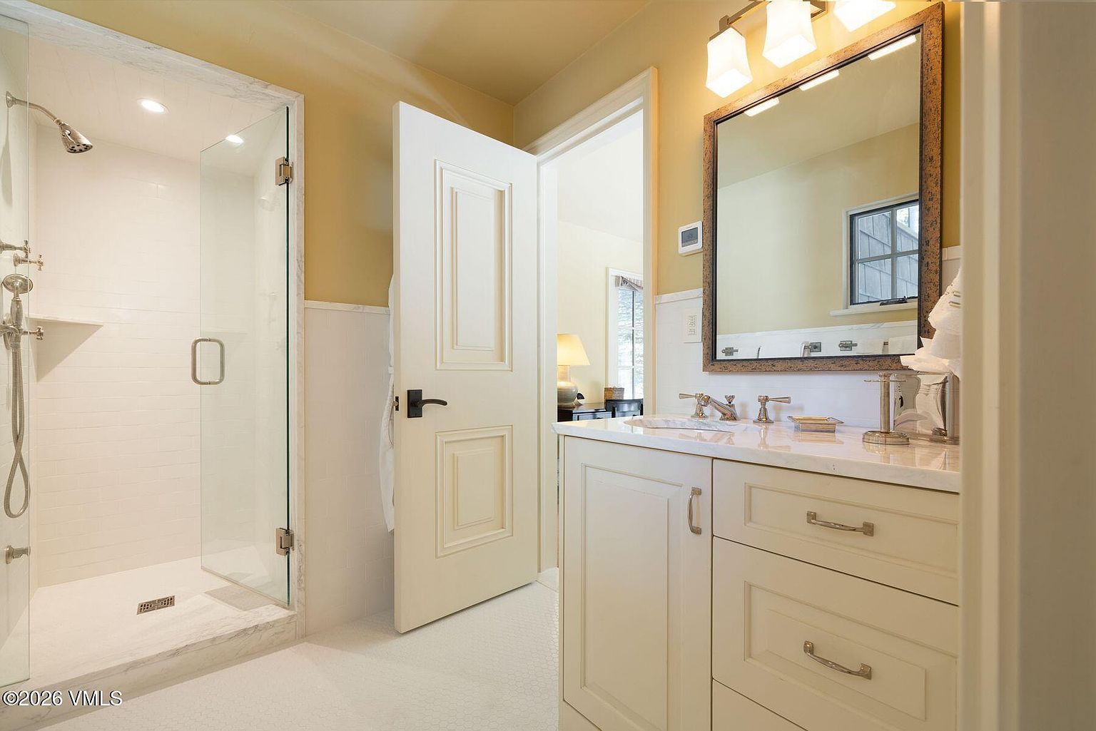 This is a well-lit bathroom featuring a glass-enclosed shower with white subway tile, a white vanity with a marble countertop, and a framed mirror with decorative lighting. The walls are painted a soft yellow, and the floor is tiled in a small white mosaic pattern. An open door leads to another room, creating a sense of spaciousness.