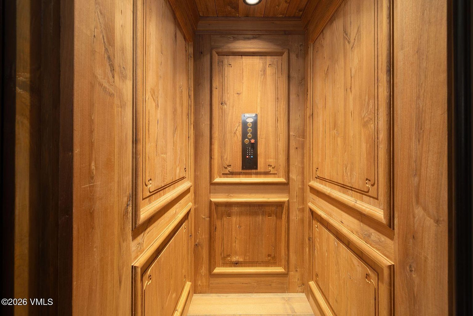 This is an interior shot of a wood-paneled elevator. The walls are covered in detailed wooden panels, and a control panel is centrally located on the back wall. The overall impression is one of luxury and craftsmanship.