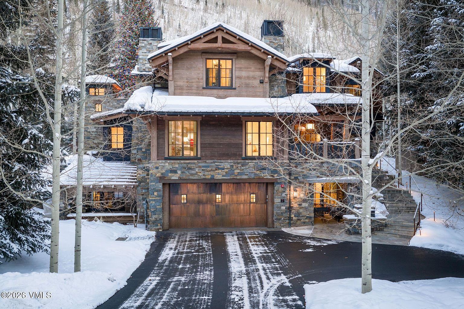 This is a front exterior view of a luxurious mountain home covered in snow. The house features a combination of stone and wood siding, with large windows providing warm interior light. A wide driveway leads to a wooden garage door, and bare trees frame the house, enhancing the wintery aesthetic.