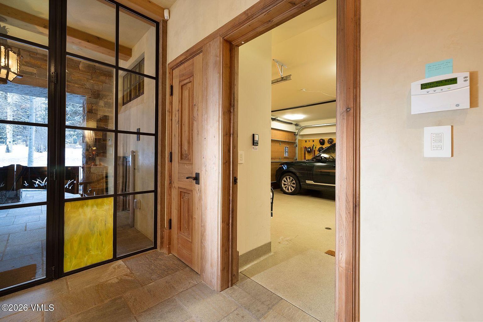 This interior shot showcases a hallway leading to a garage. The hallway features a wooden door and trim, contrasting with the light-colored walls. A glimpse into the garage reveals a luxury vehicle, suggesting high-end living. The flooring appears to be stone tile, adding a touch of elegance.