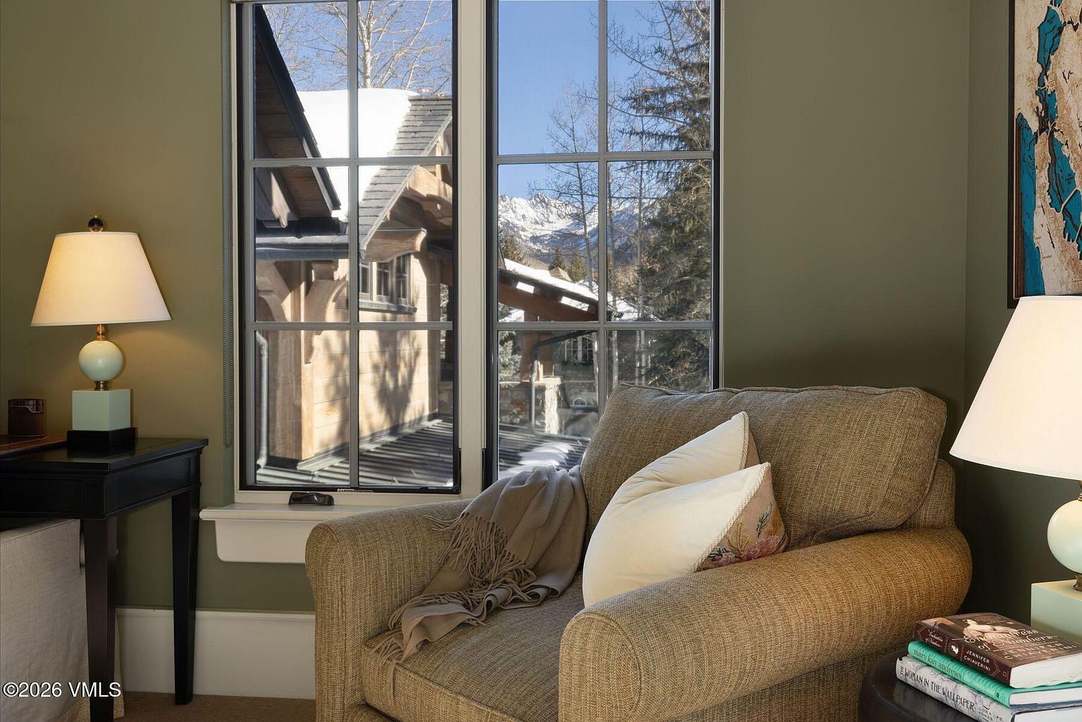 This is a cozy living room featuring a comfortable armchair with a throw blanket and pillows, positioned near a window with a view of snow-covered rooftops and mountains. A lamp sits on a side table next to the chair, creating a warm and inviting atmosphere. The room is painted in a muted green tone, enhancing the sense of relaxation and comfort.