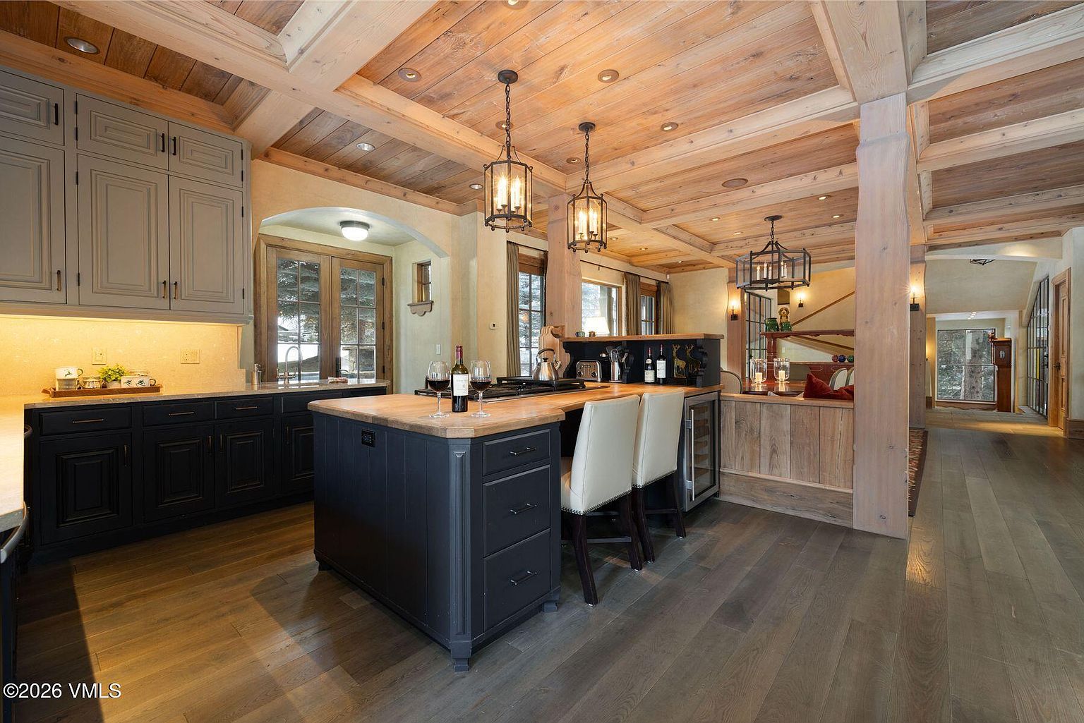 This is a warm and inviting kitchen featuring a wood-paneled ceiling with exposed beams and pendant lighting. The kitchen island has a butcher block countertop and dark cabinetry, complemented by lighter gray cabinets along the wall. The space includes modern appliances and a wine fridge, creating a blend of rustic charm and contemporary convenience.