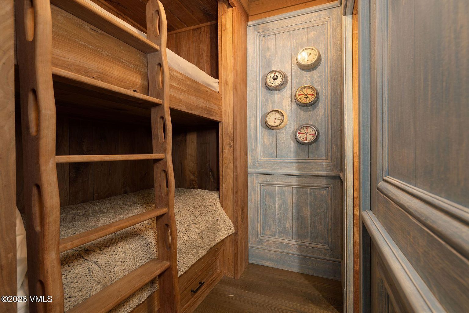 This image showcases a cozy bedroom featuring custom-built wooden bunk beds with a ladder. The beds are neatly made with textured blankets, and a set of decorative clocks adorns the adjacent blue-painted wall, adding a whimsical touch. The room exudes a rustic and charming atmosphere, perfect for a guest room or children's bedroom.
