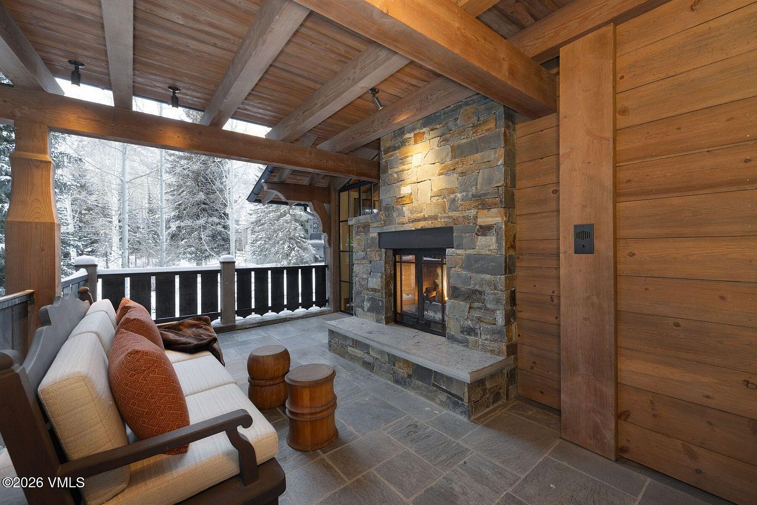 This image showcases a cozy outdoor patio or balcony area, featuring a stone fireplace as a focal point. Comfortable seating is arranged to enjoy the warmth of the fire and the snowy view beyond the railing. The space is enclosed with wooden walls and a wooden ceiling, creating a rustic and inviting atmosphere.