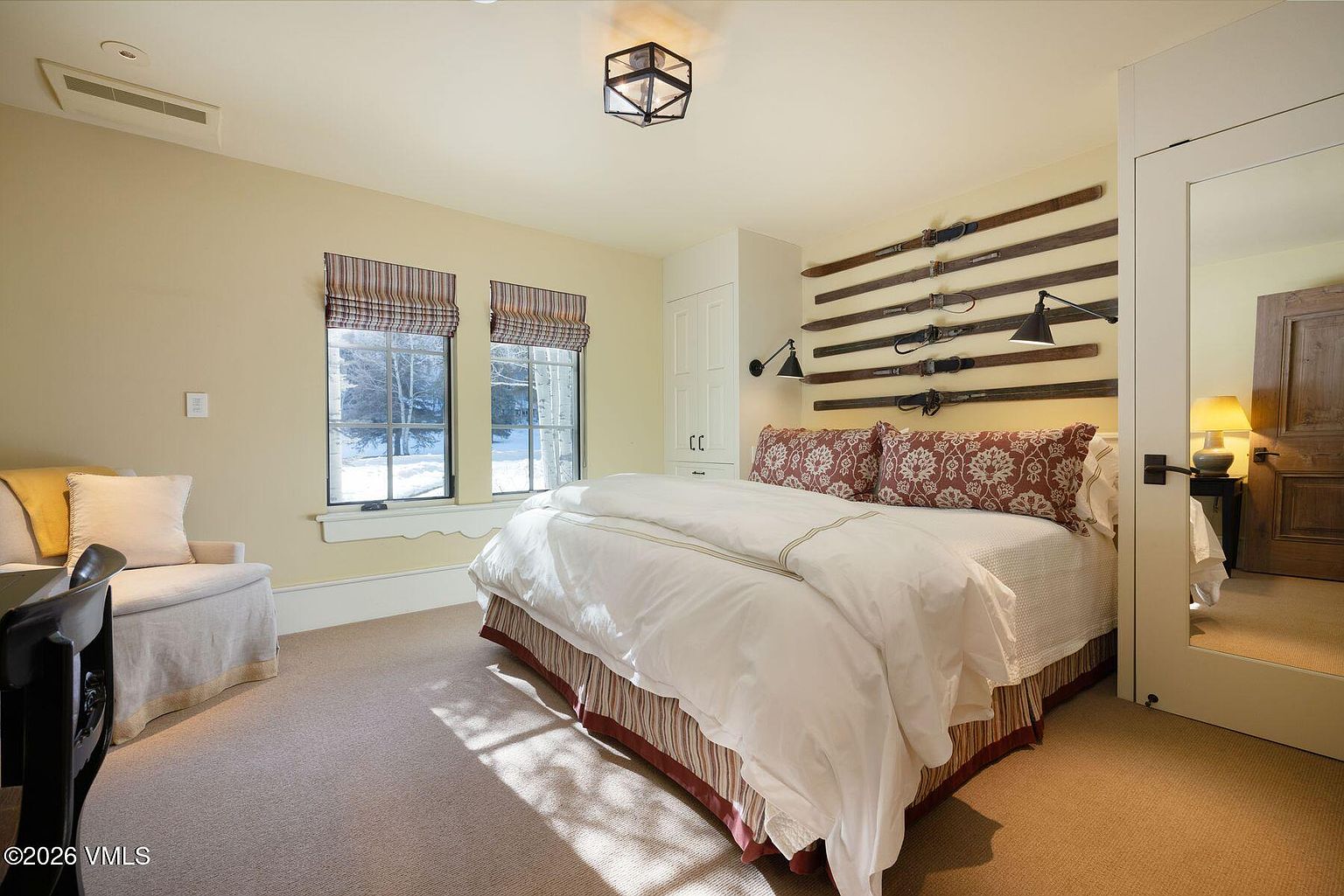 This is a cozy bedroom featuring a bed with white linens and decorative pillows, flanked by unique wall-mounted ski decor and sconce lighting. The room has two windows with striped blinds, a neutral color scheme, and carpet flooring, creating a warm and inviting atmosphere. A glimpse of an adjacent room is visible through a mirrored door.